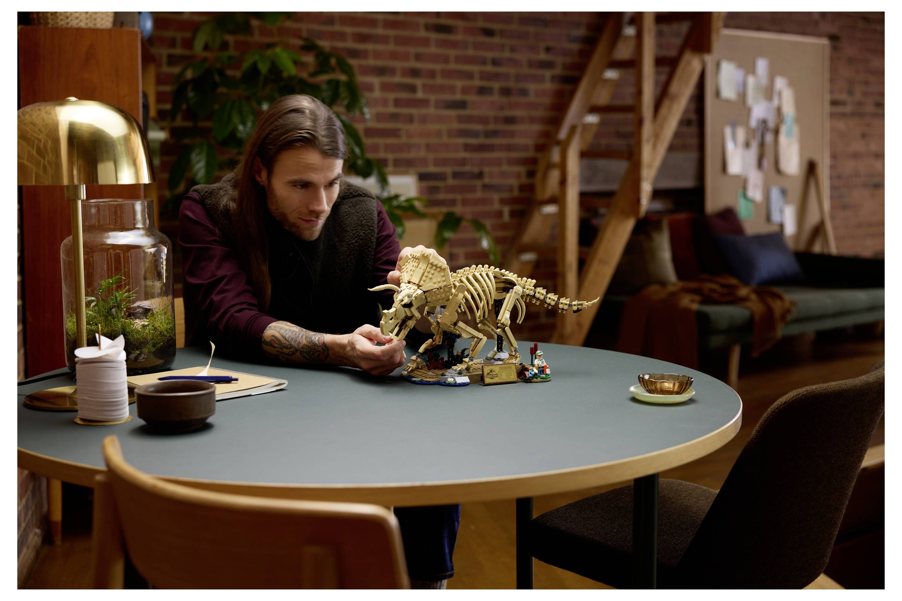Muž studuje model kostry dinosaura na kulatém stole v útulném studiu, v jeho blízkosti jsou další LEGO dílky.