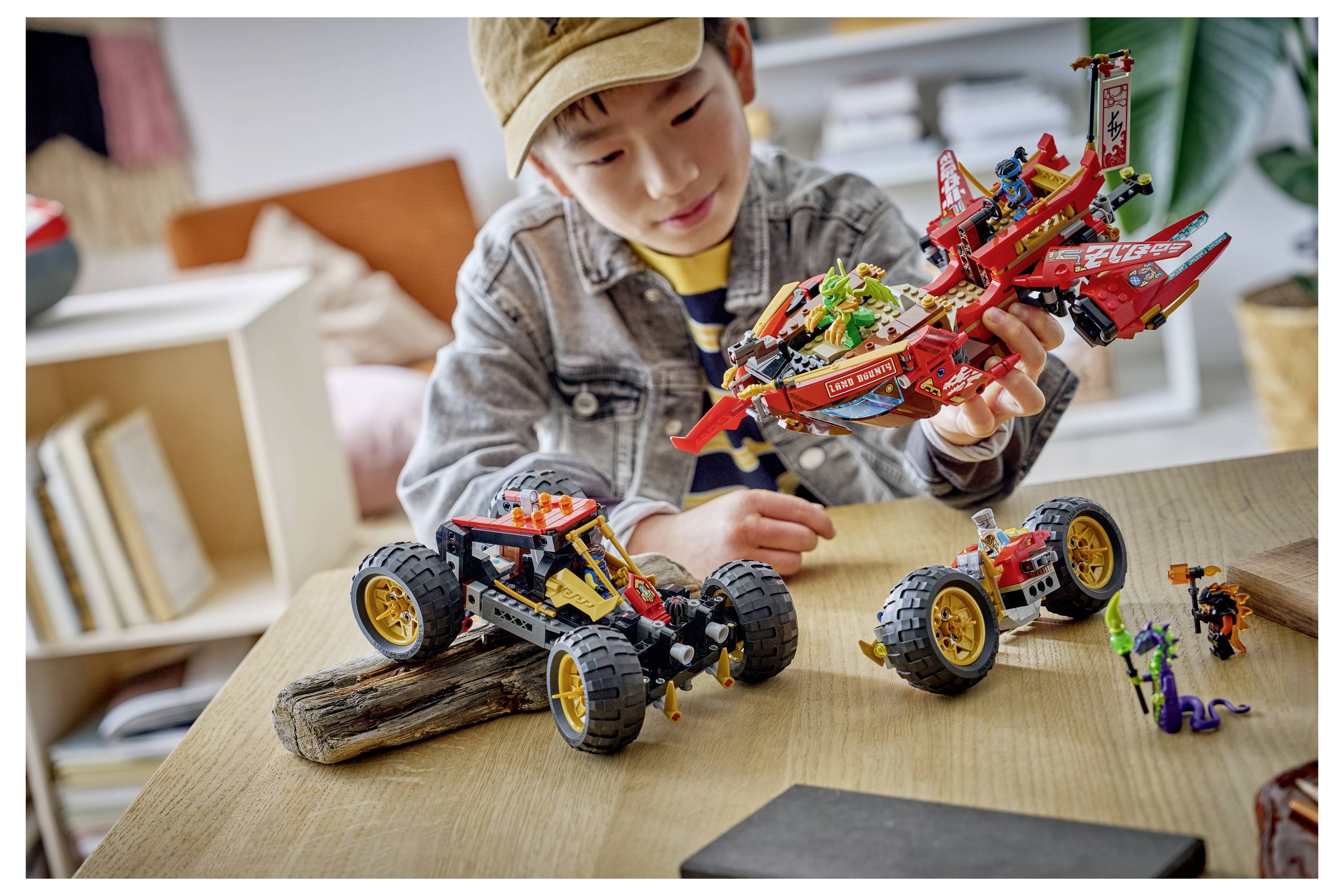 Dítě si prohlíží červený letadlový model v LEGO stylu na stole s dvěma malými hračkovými vozidly před sebou.