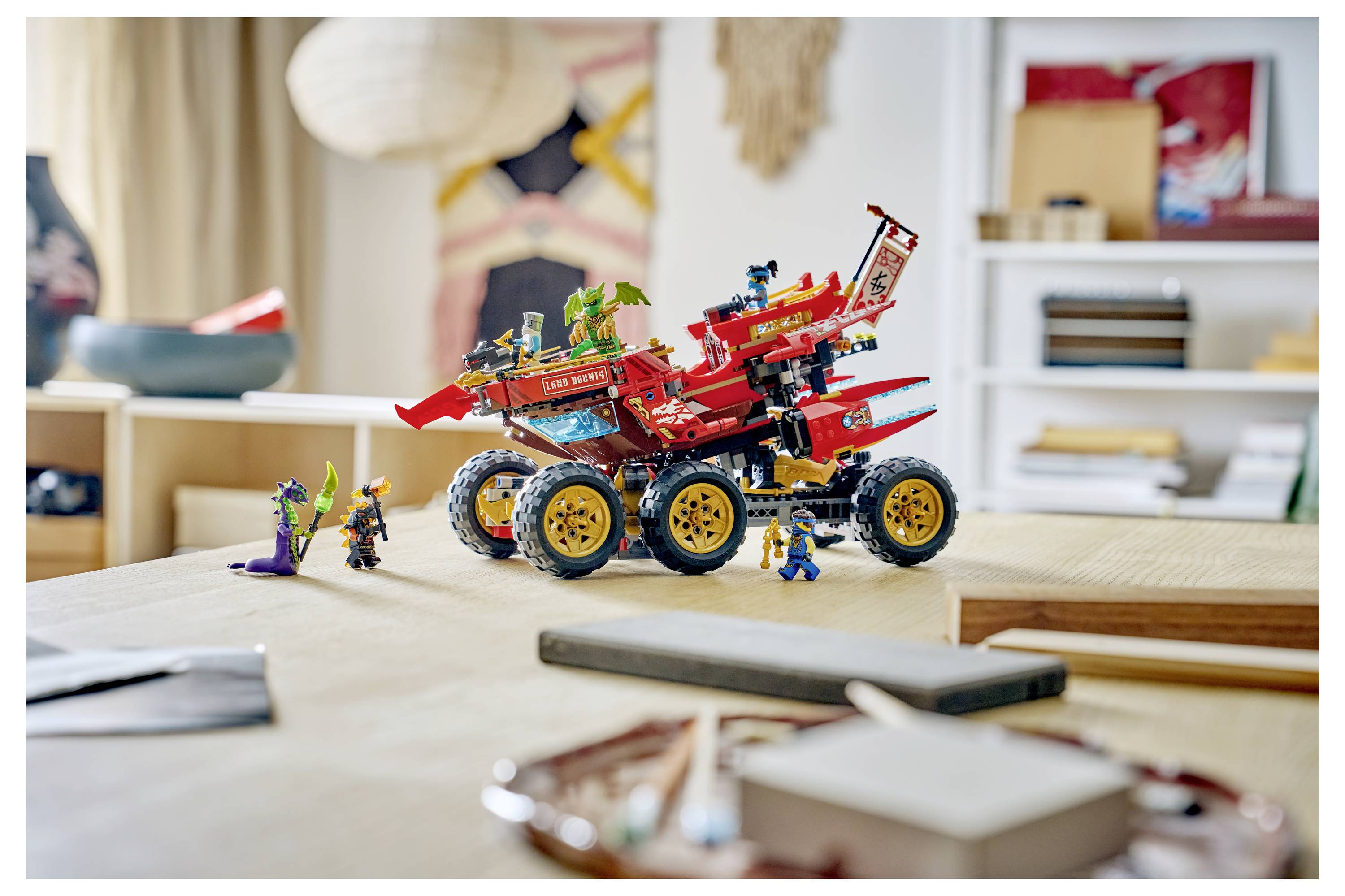 Červené vozidlo v LEGO stylu určené pro terénní jízdu s minifigurkami sedí na stole v obývacím pokoji, zobrazeno v detailním záběru vnitřního scénáře.