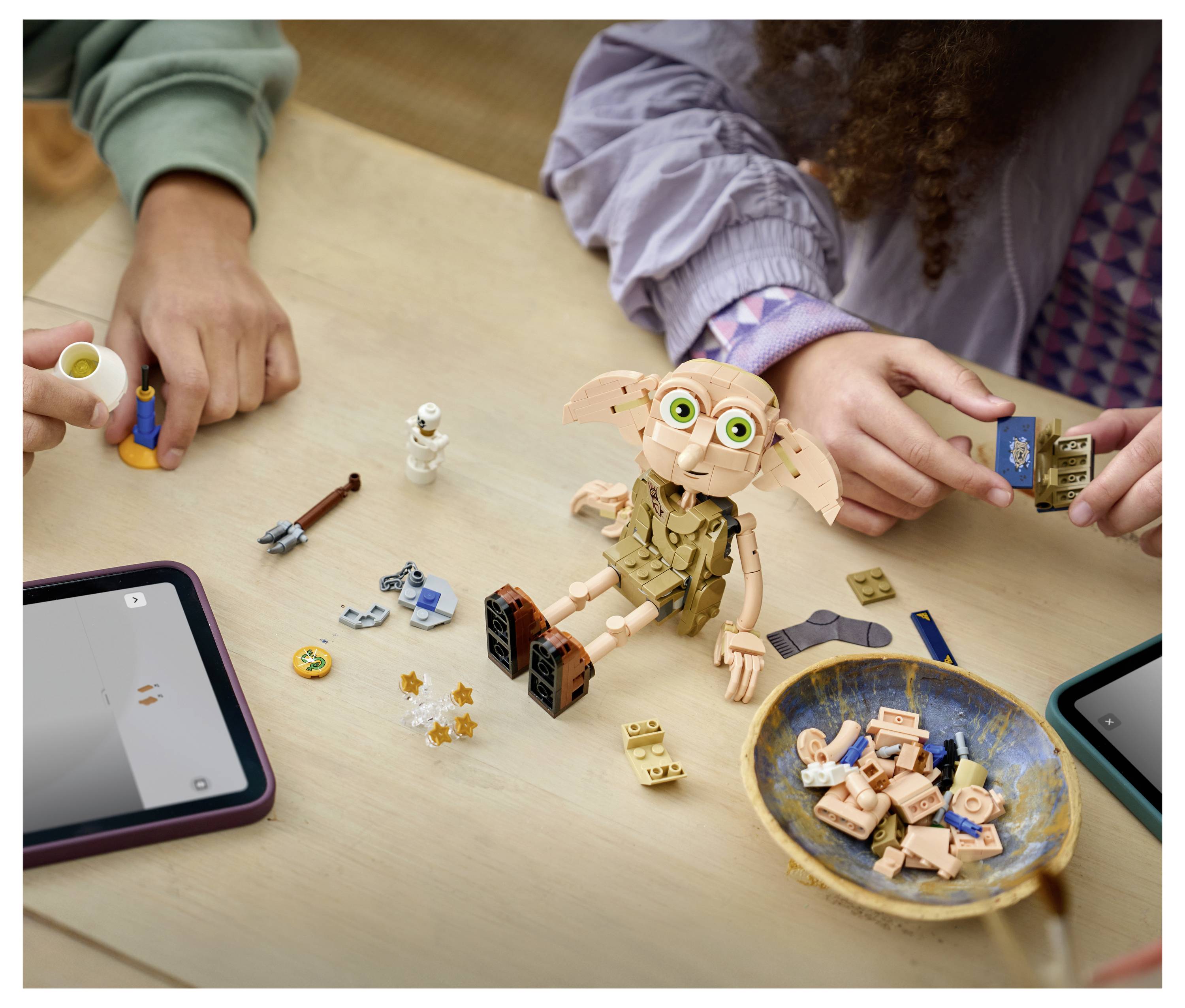 Děti skládají LEGO-style stavbu Dobbyho na stole, s rozptýlenými kusy, tablety a miskou součástek poblíž.