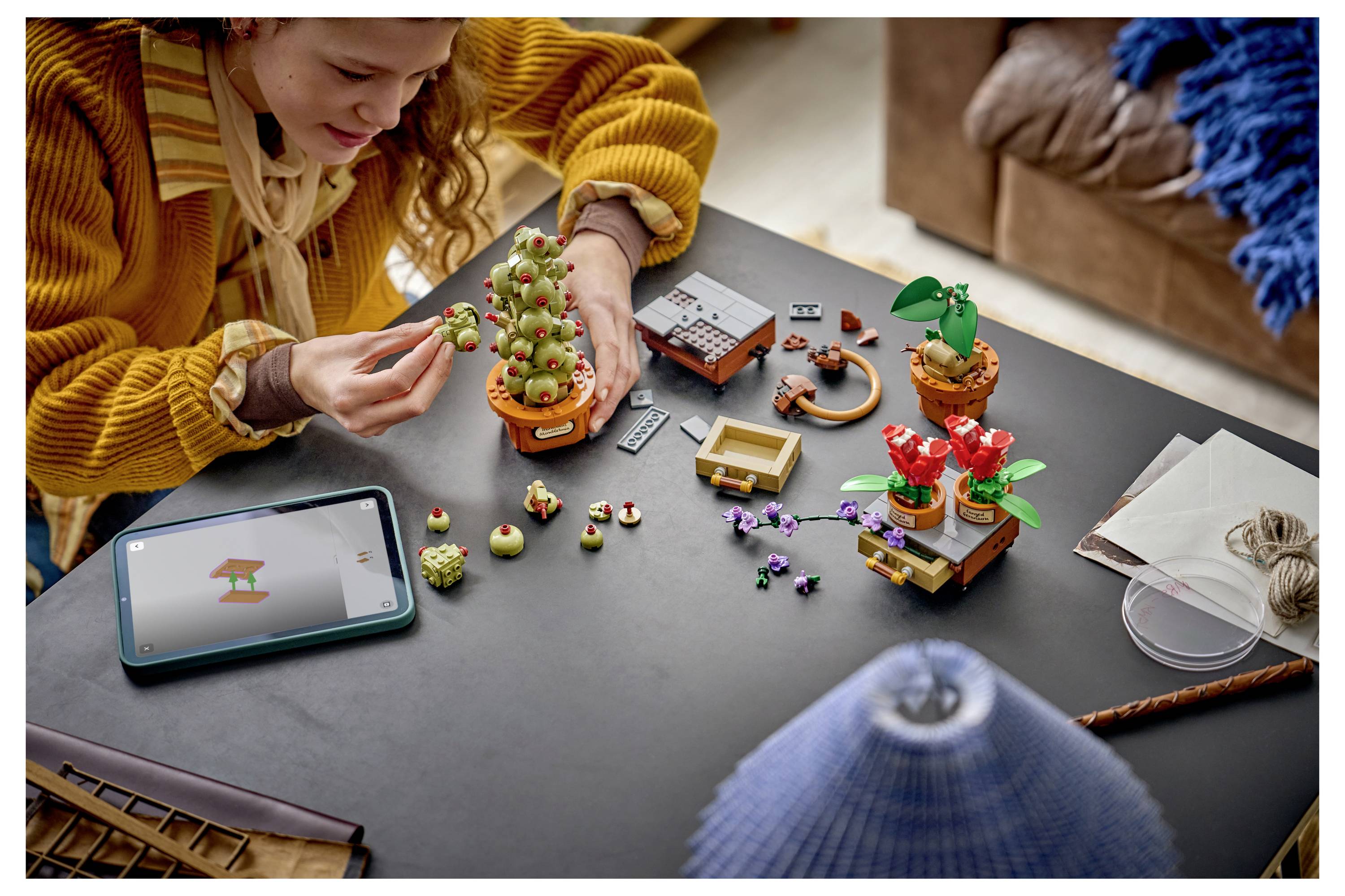 Osoba skládající malé растениové modely podobné LEGO u stolu, vedle ní tablet zobrazující pokyny.