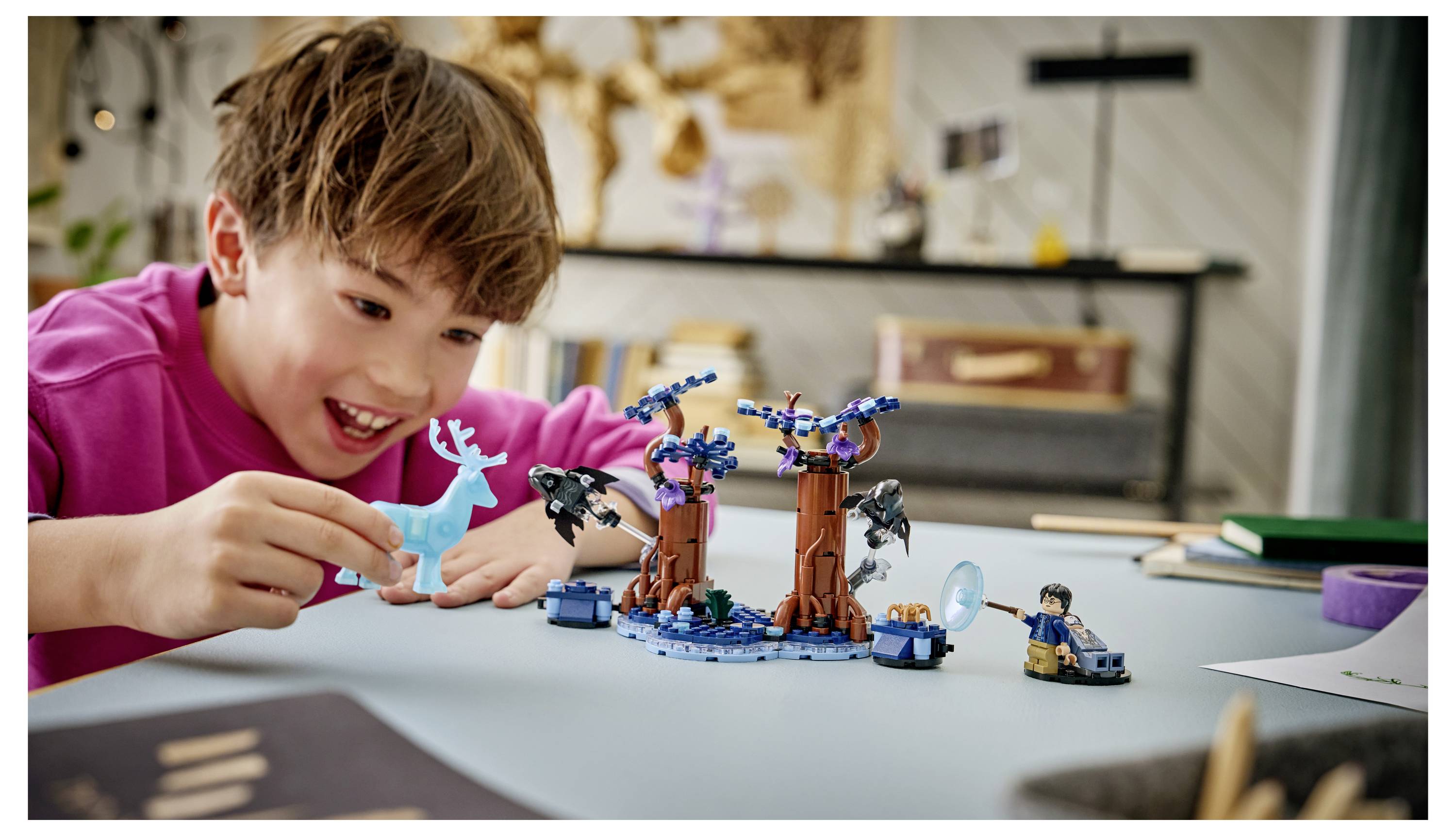 Chlapec v růžové košili se usmívá, zatímco si hraje se stavebnicí ve stylu LEGO s fantastickým stromem a modrými figurkami u stolu.