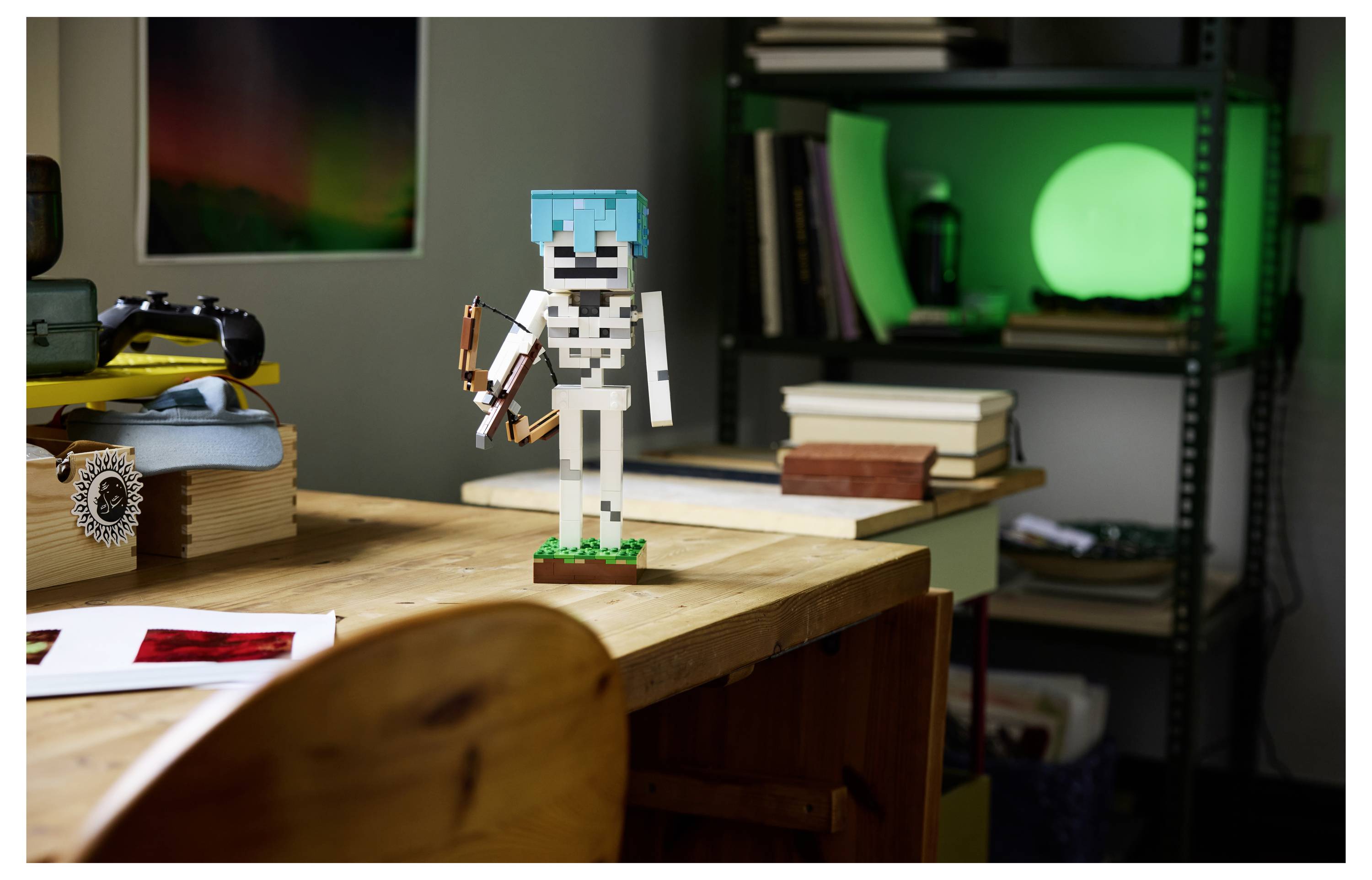 Malá LEGO postavička kostlivce s modrým přilbou stojí na dřevěném stole vedle knih v místnosti se zářící zelenou lampou.