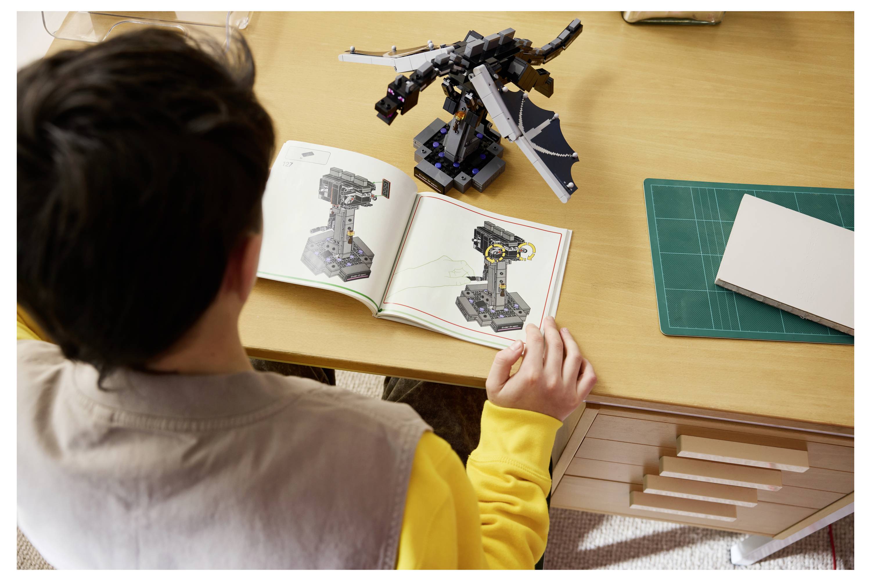 Osoba stavící model v LEGO stylu u psacího stolu, která porovnává sestavený drak podobný předmět s pokyny v otevřeném manuálu.