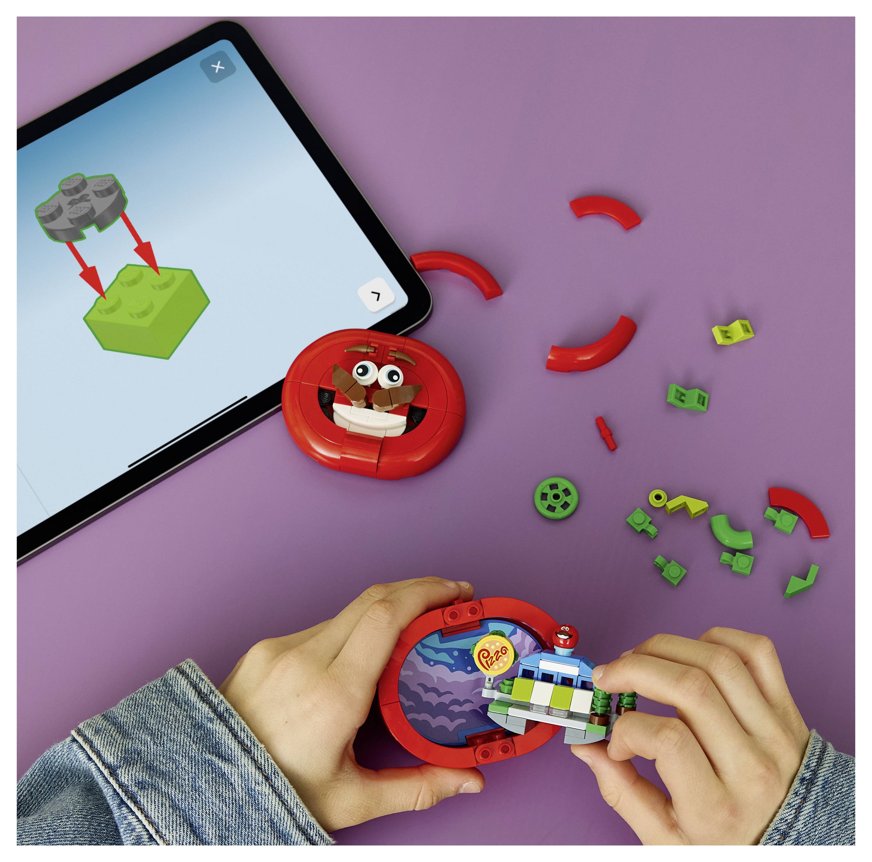 Ruce skládají červenou kruhovou hračku z malých kusů podobných LEGO, zatímco tablet zobrazuje návod na montáž na fialovém pozadí.