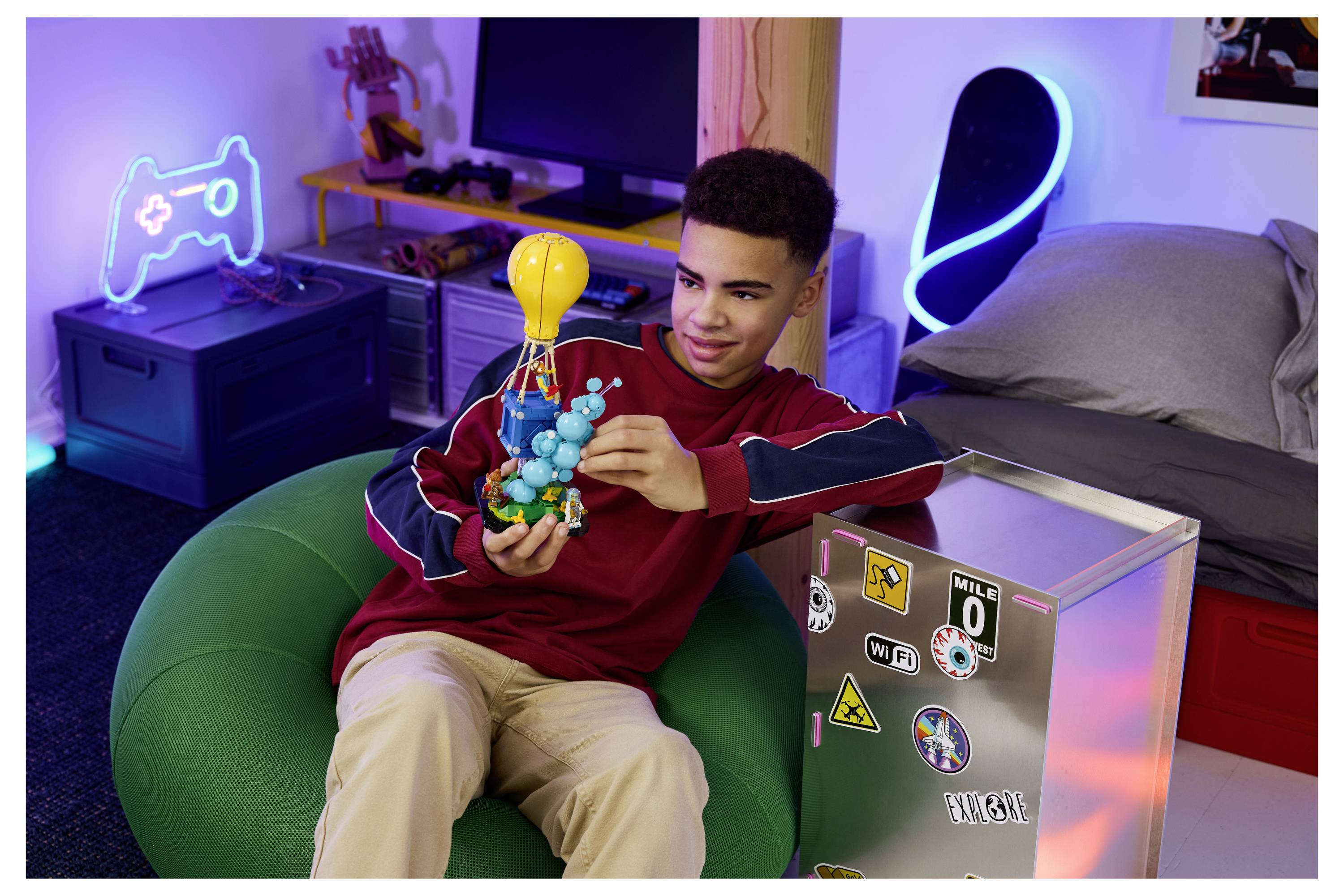 Teenagerský chlapec odpočívající v herním setup připomínajícím ložnici, sestavující barevný molekulární model mientras sedí v zelené pytlíkovací židli.