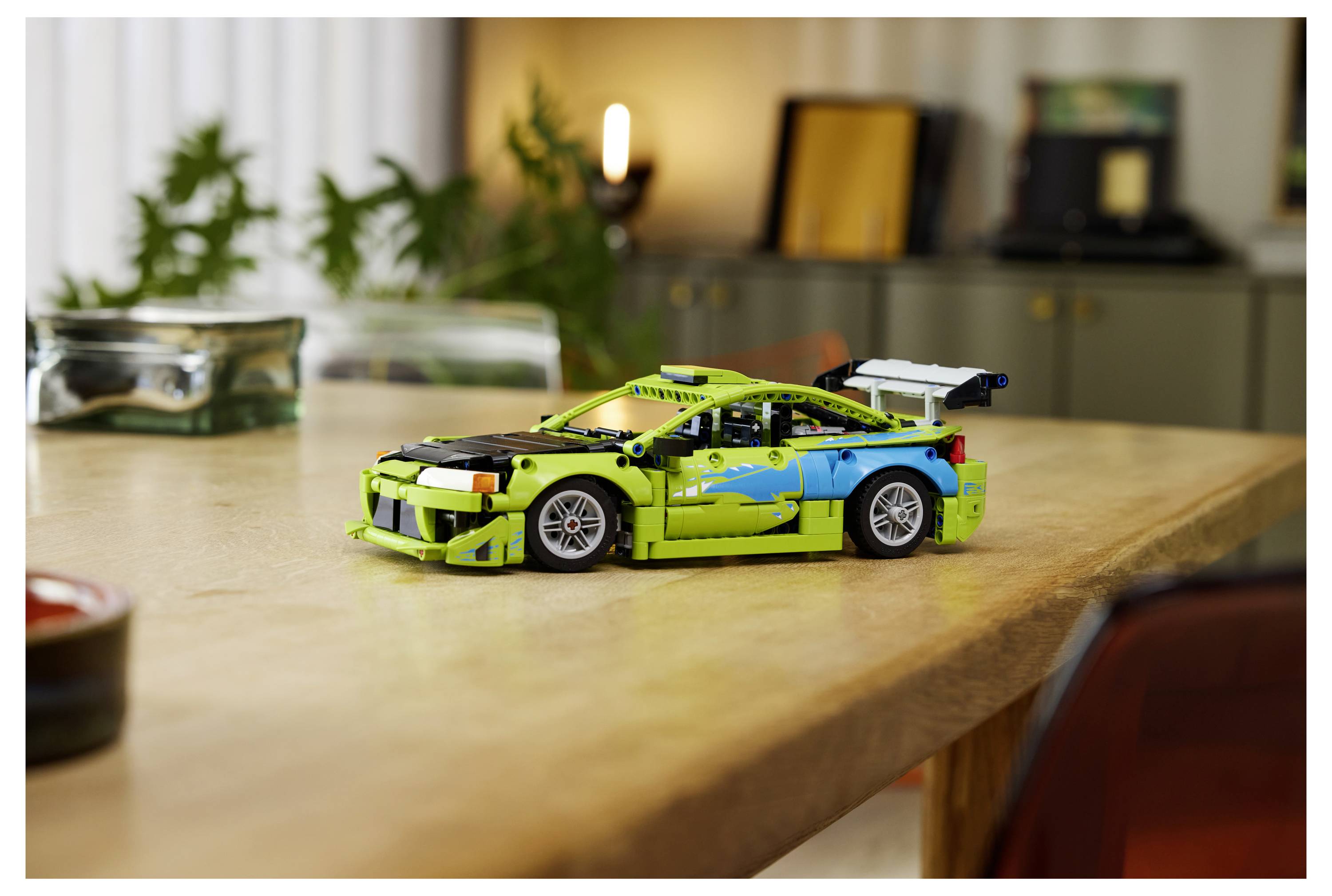 Jasně zeleno-modrý model závodního auta LEGO sedí na dřevěném stole v měkce rozmazaném interiéru místnosti.