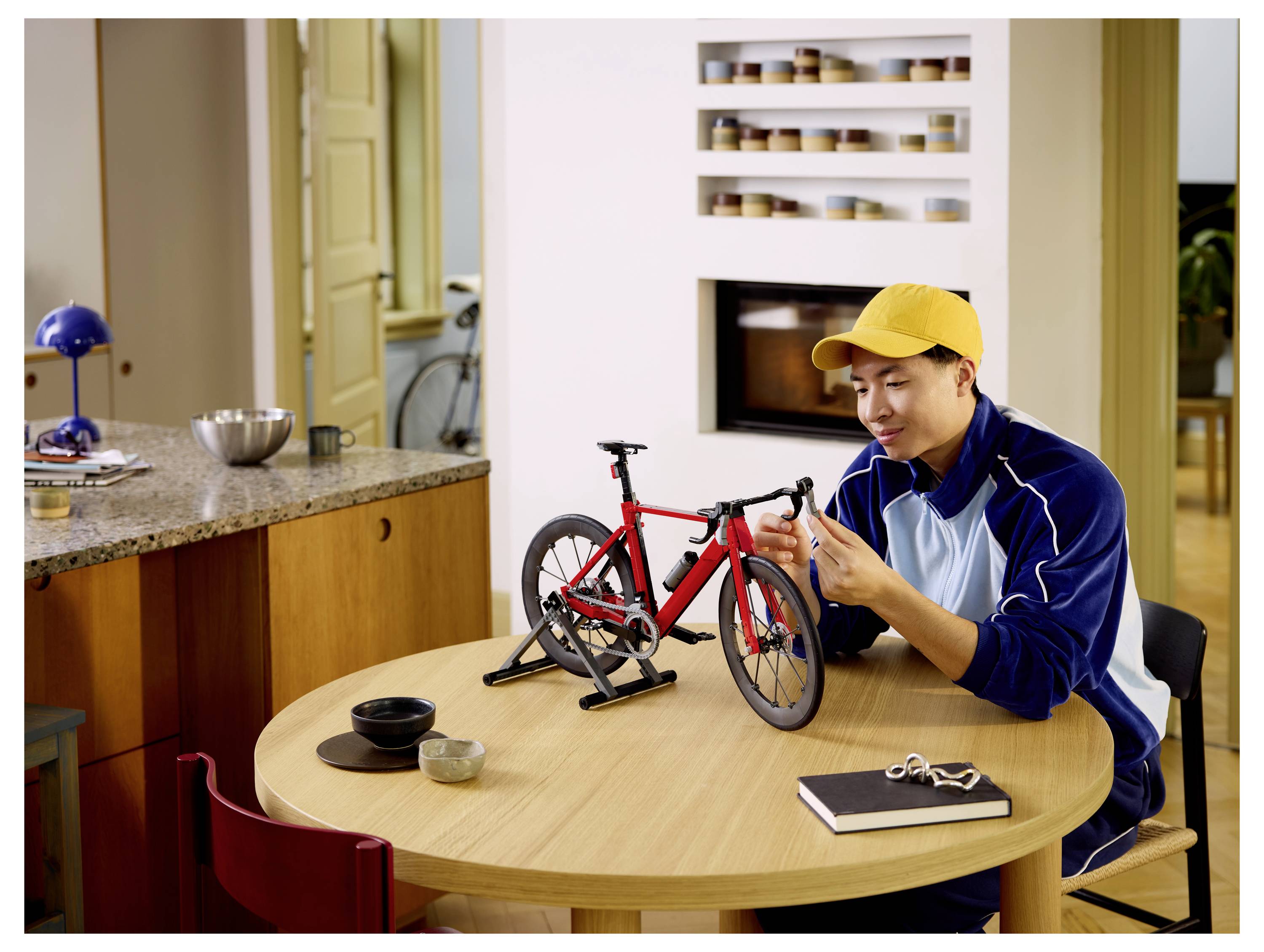 Muž v žlutém klobouku upravuje malý červený model bicyklu na jedálně v jasné kuchyni.