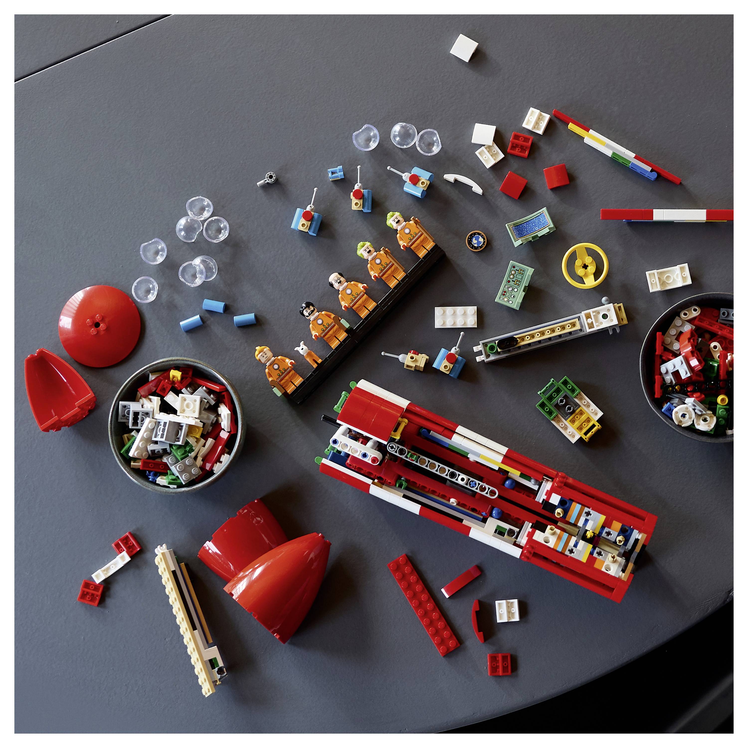 Rozházené LEGO kostičky a částečné červené modely na šedém stole, včetně koleček, cihel, minifigurek a kulatých kontejnerů s různorodými součástkami.