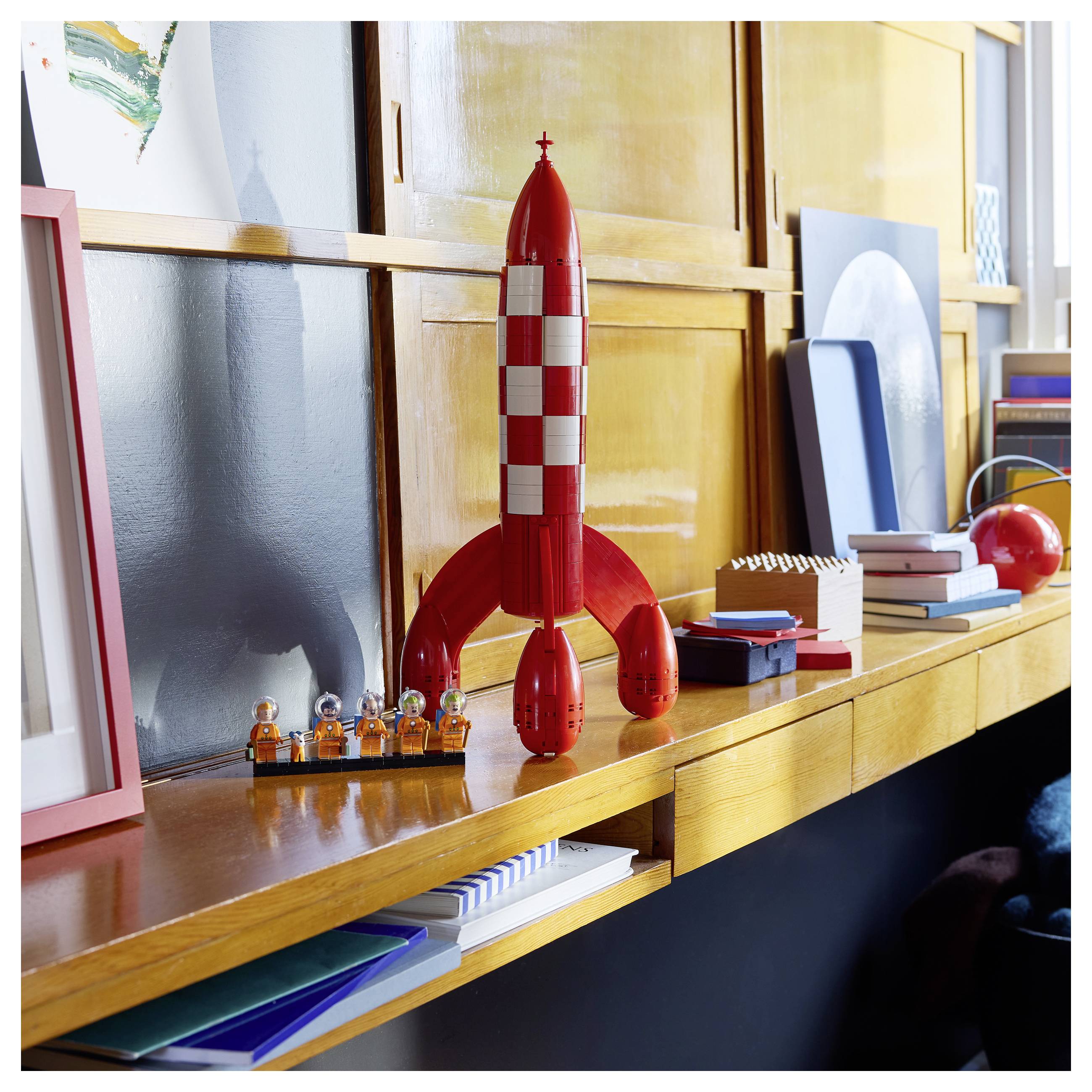 Červená hračka raketa na dřevěné polici, s malými figurkami astronautů a knihami, v jasně osvětlené místnosti.