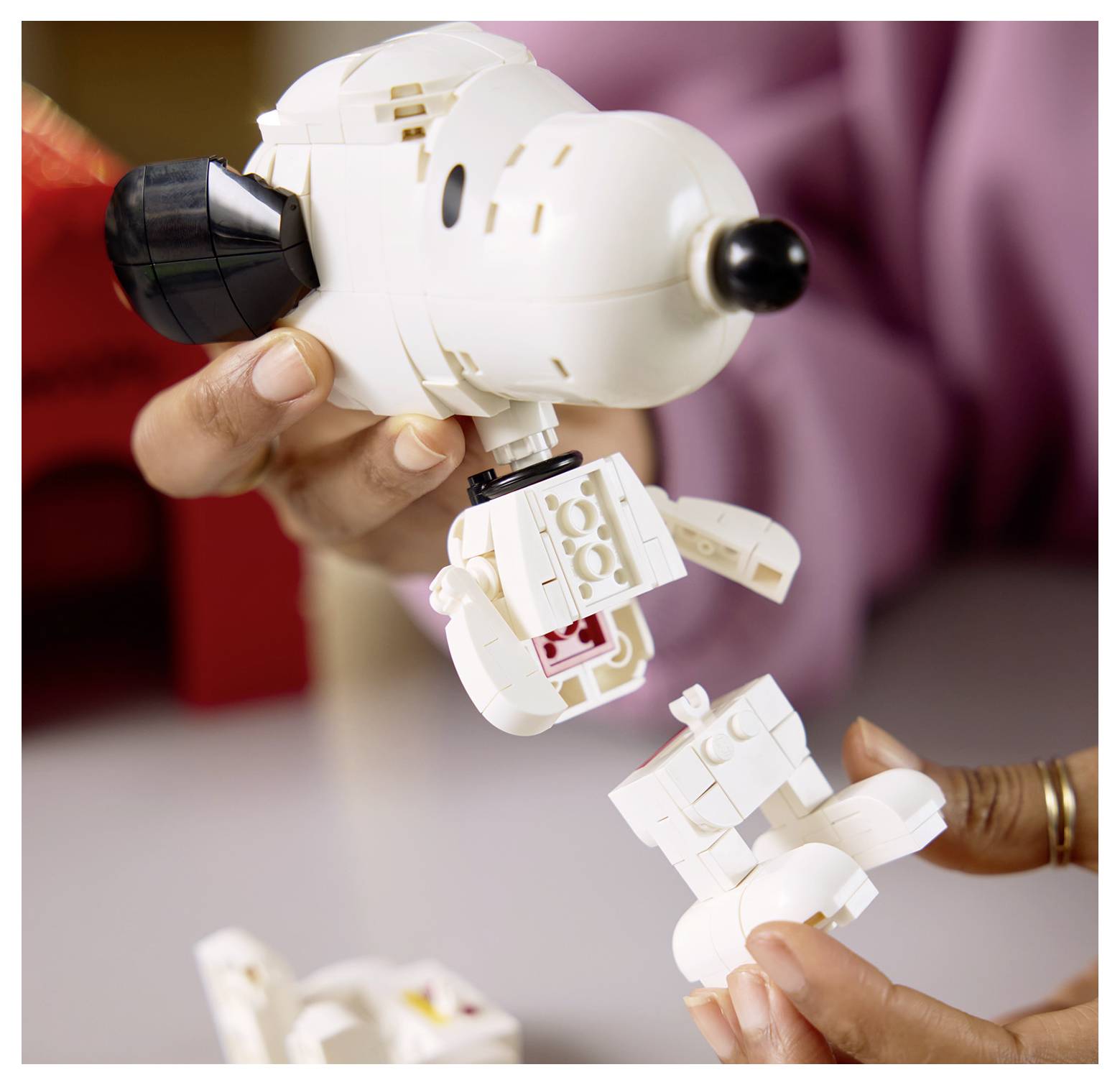 Ruce držící bílou figurku LEGO Snoopyho, která se montuje, s viditelnou rozloženými součástkami.