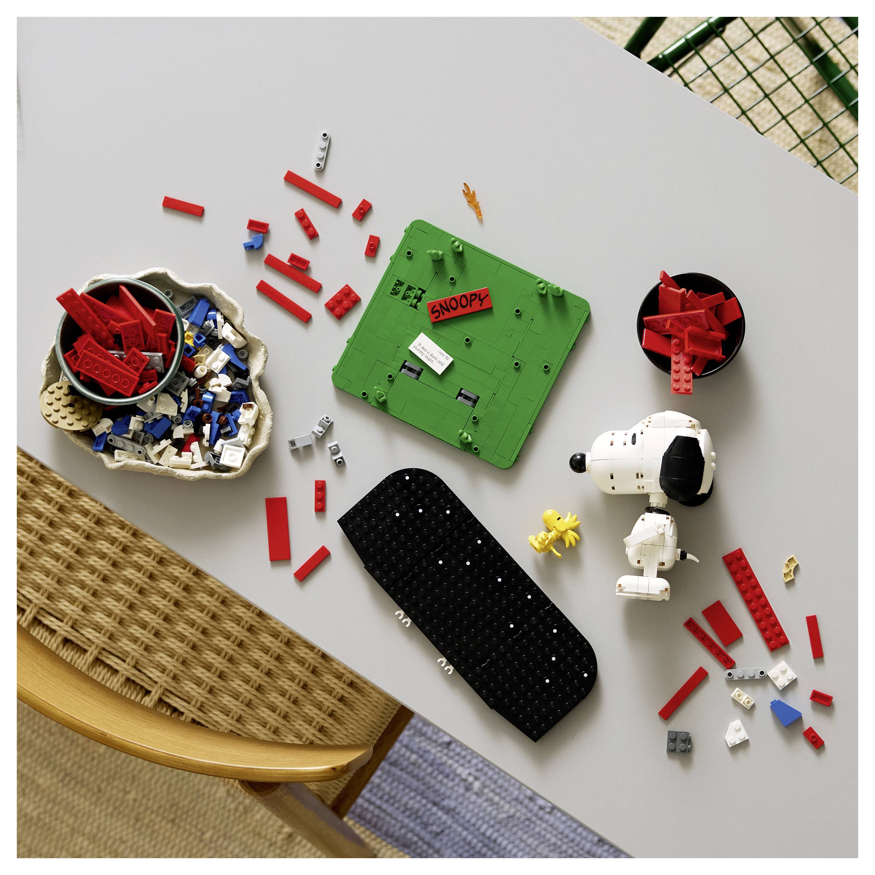 Pohled zeshora na rozptýlené LEGO součástky s motivem Snoopyho a pokyny na stole, s červenými, bílými, černými a zelenými kostičkami a hotovou figurkou Snoopyho