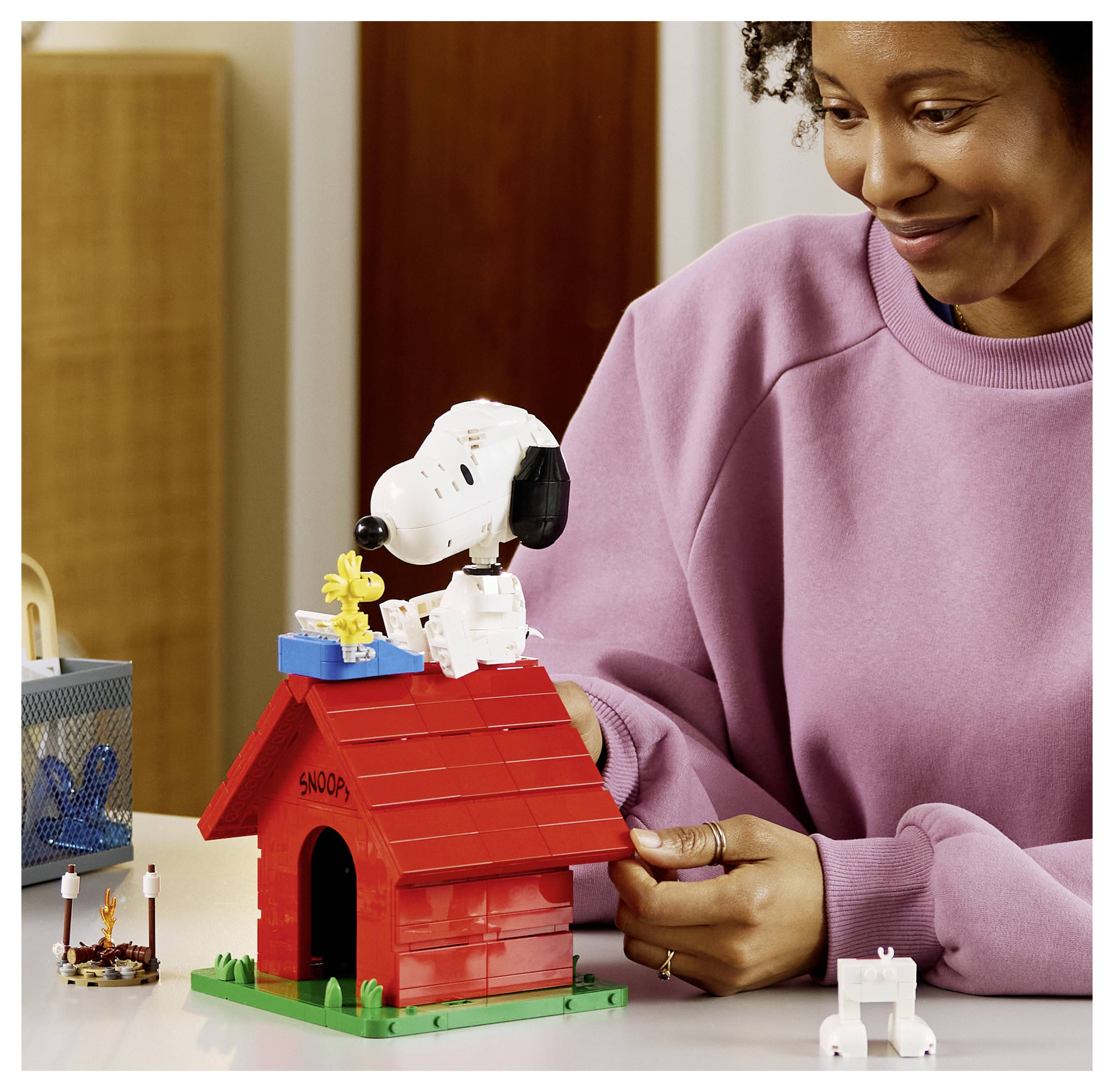 Žena se usmívá při stavbě červeného legoového psího boudy se figurkou Snoopyho nahoře, s malou figurkou bílého psa poblíž.