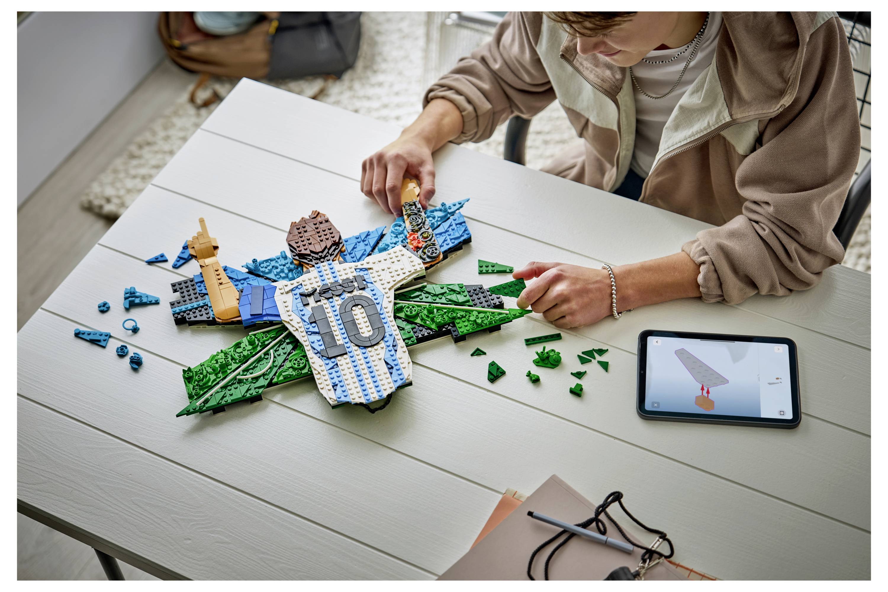 Osoba skládající letadlo LEGO na stole, umisťující modré a zelené díly kolem dresu podobného středu označeného číslem 10, s tabletem zobrazujícím poblíž raketu.