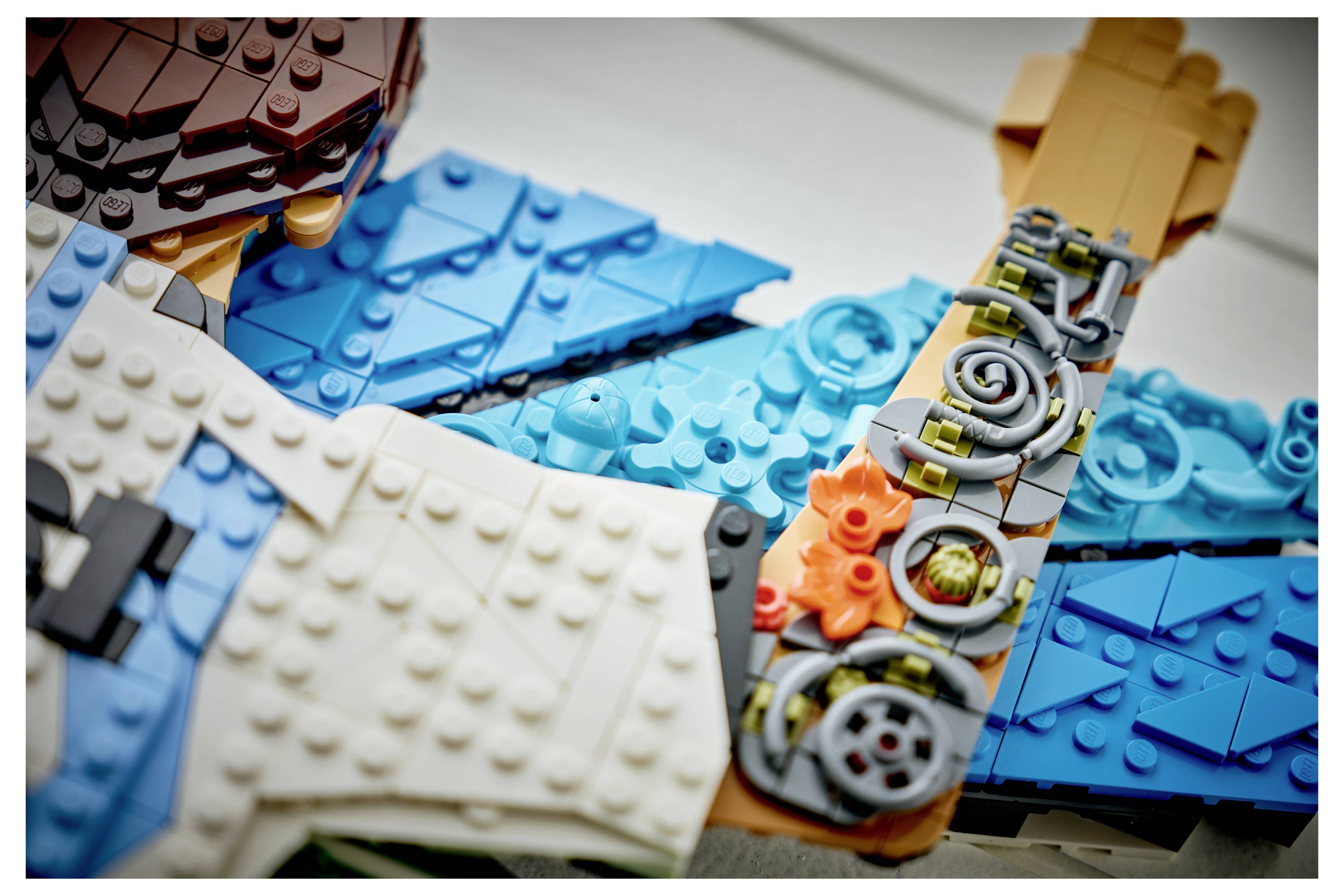 Detailní snímek různorodých LEGO dílků a součástí minifigurek rozložených na stole, které zobrazují modré, bílé, hnědé a oranžové kostky a ozubená kola.