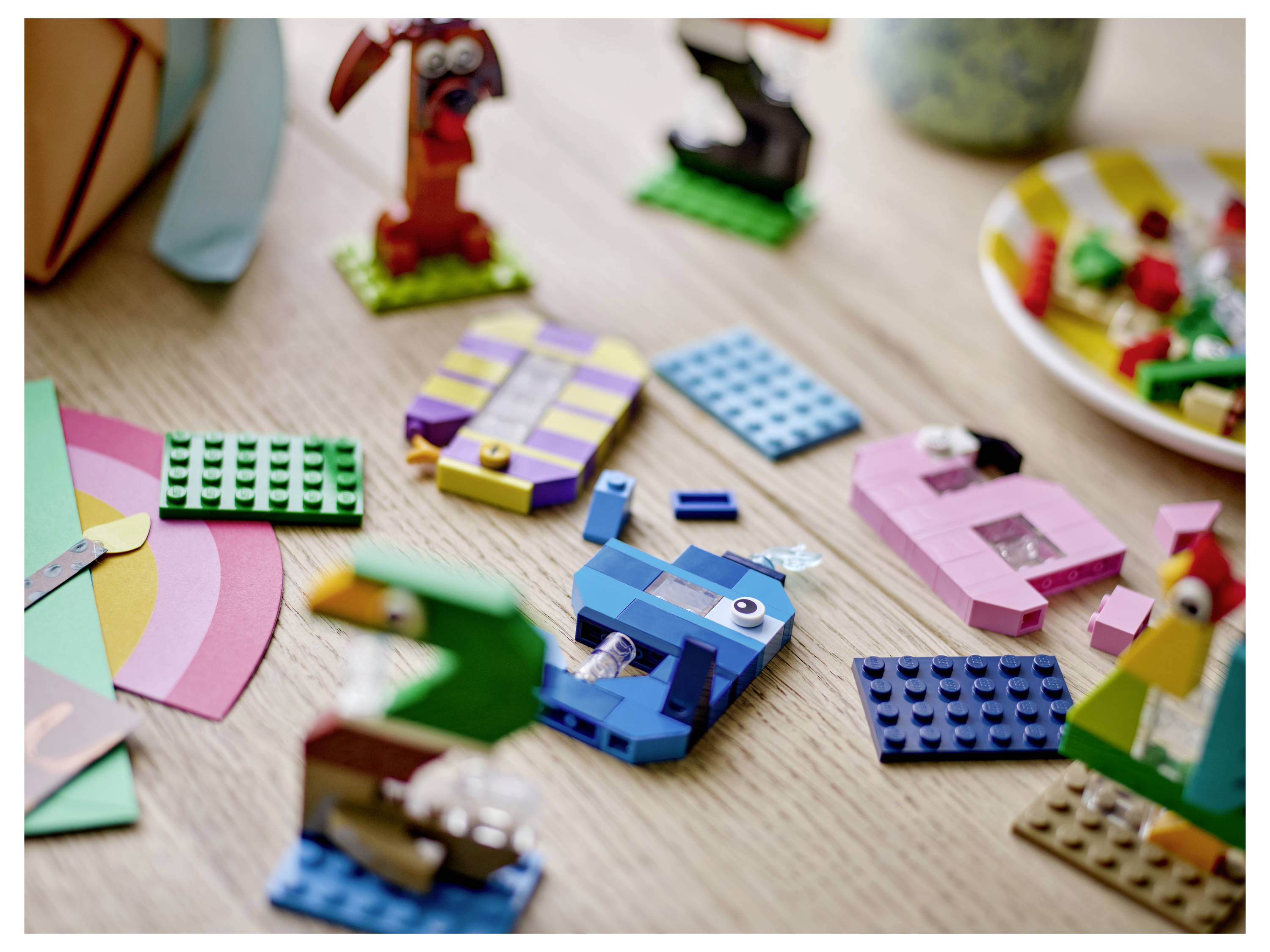 Různobarevné LEGO kostky a malé stavby rozptýlené na dřevěném stole, uprostřed modrá hranaté hračkové vozidlo.