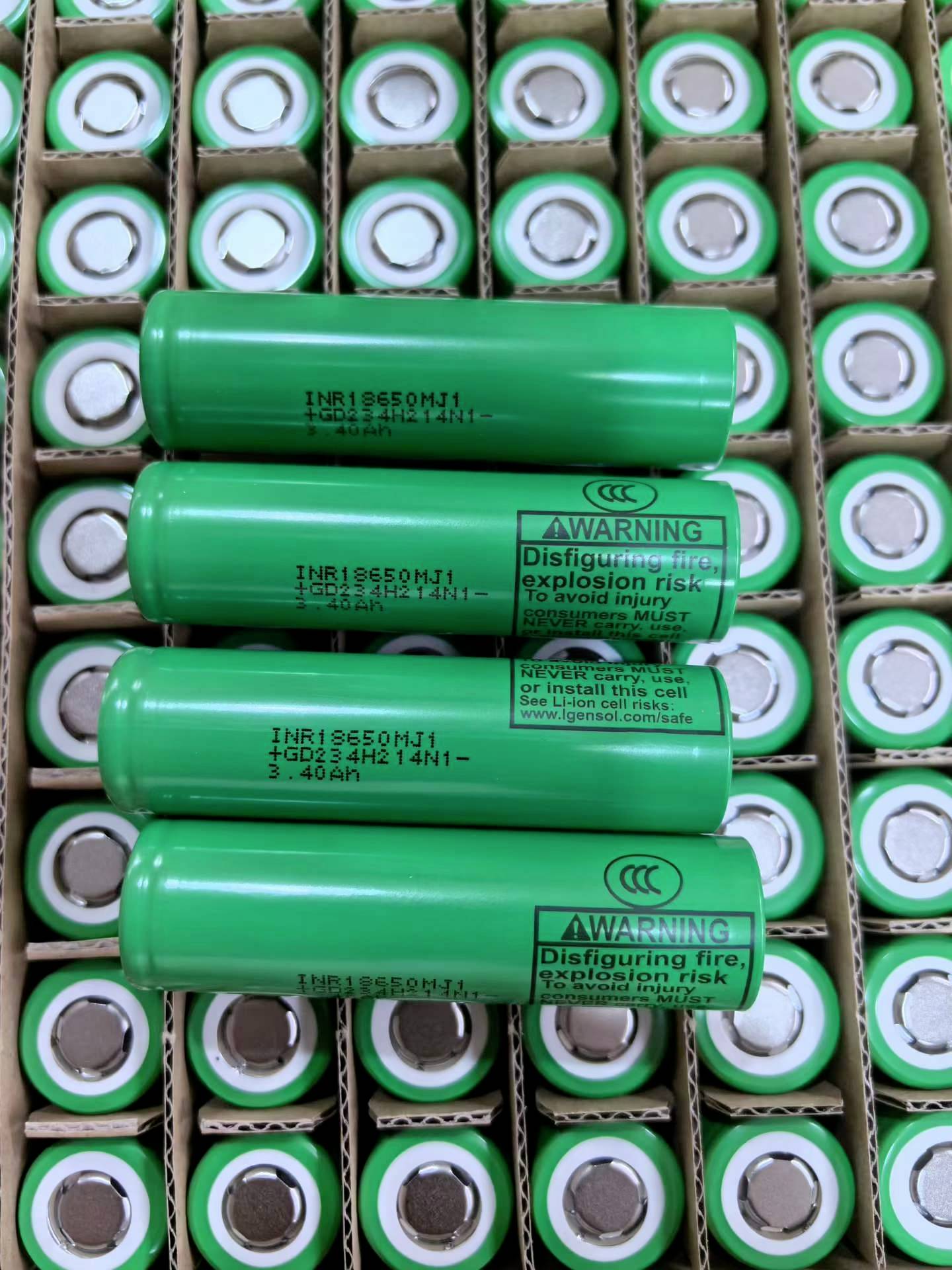 Skládané zelené lithium-ionové baterie v papírových přepravkách, s několika většími články označenými varovným textem o riziku požáru a výbuchu.