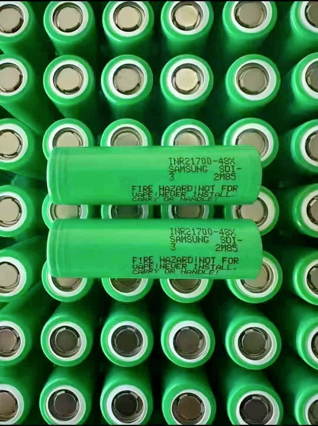Detailní záběr na zelené nabíjecí články Samsung INR21700-48G seřazené v řadách, přičemž dva články leží vodorovně přes mnoho vzpřímených článků.