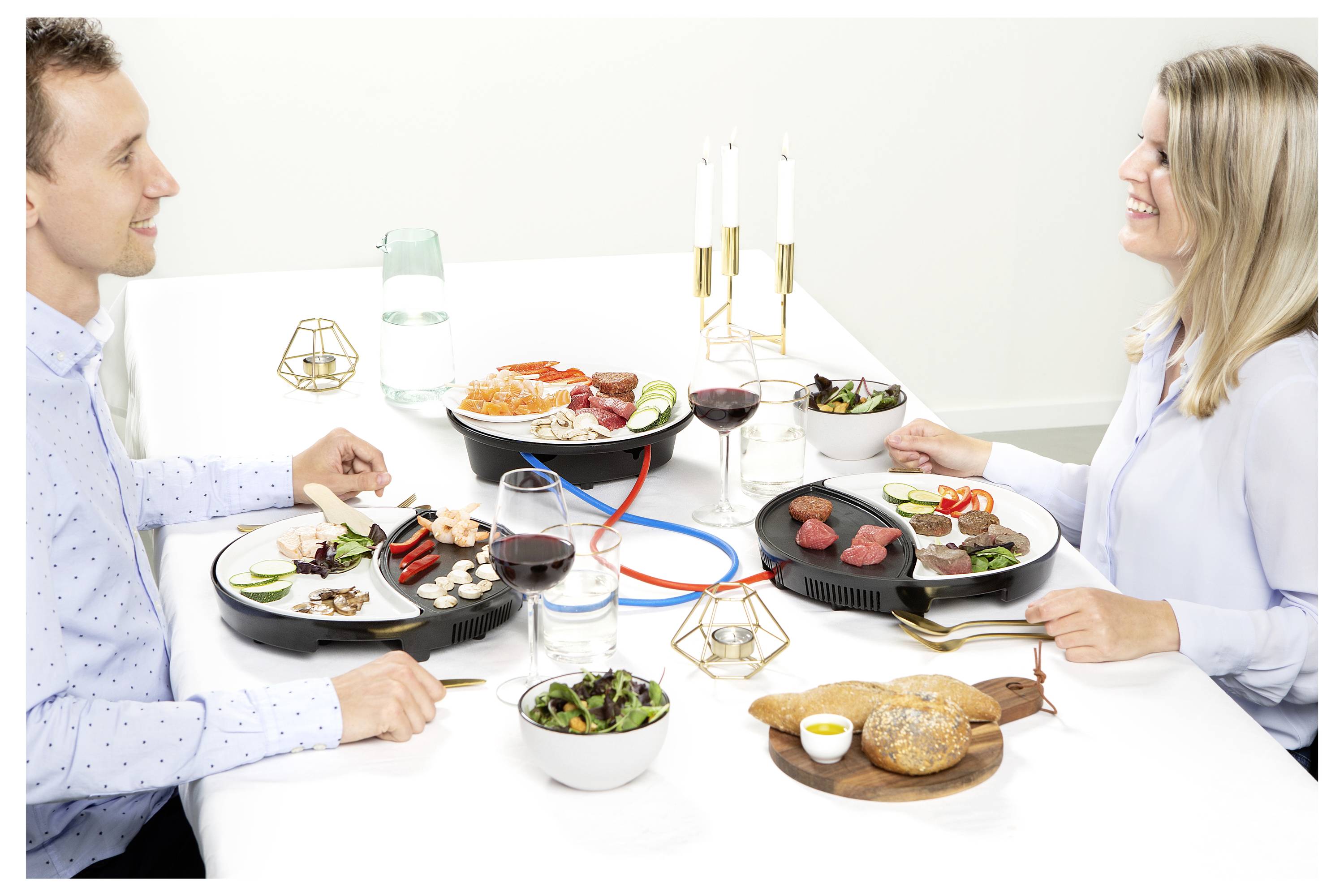 Muž a žena si dělí fondue při jídle u jídelního stolu s talíři masa, zeleniny, chleba, vína a svíček.