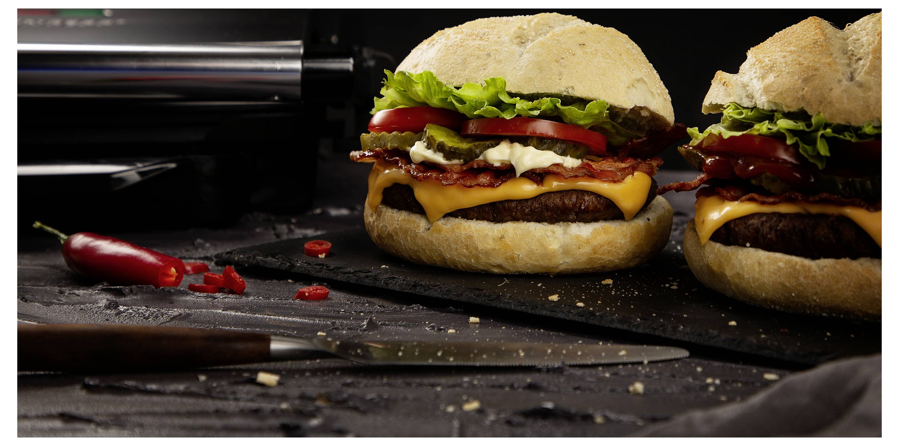 Detailní záběr na dva cheeseburgers s salátjem a rajčetem na tmavé pracovní desce, s nožem a červeným pepřem v popředí.