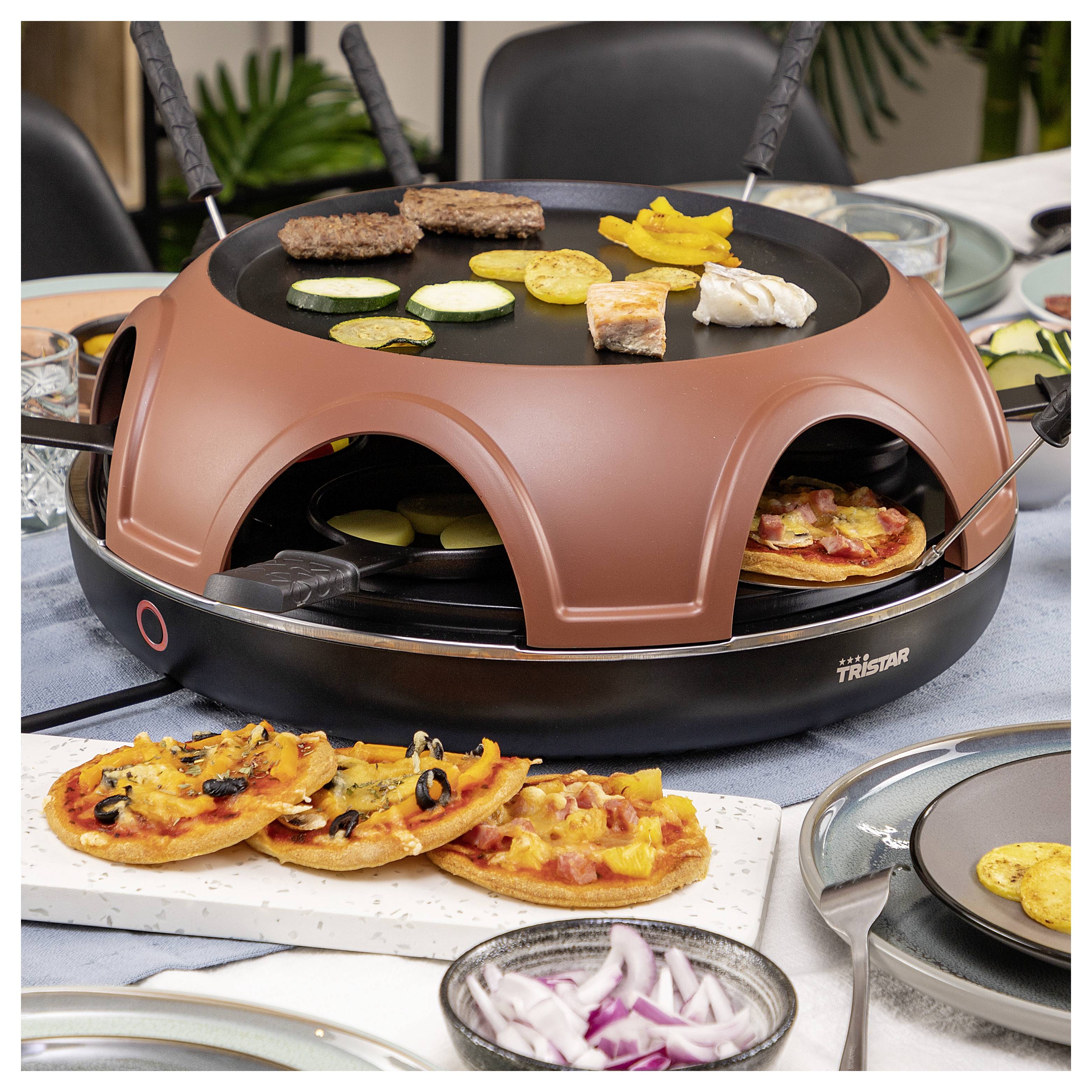 Stolní gril a zařízení na raclette s masem, zeleninou, krevetami a malými pizzami na vaření, s plátky jídla a krájené cibule kolem něj.