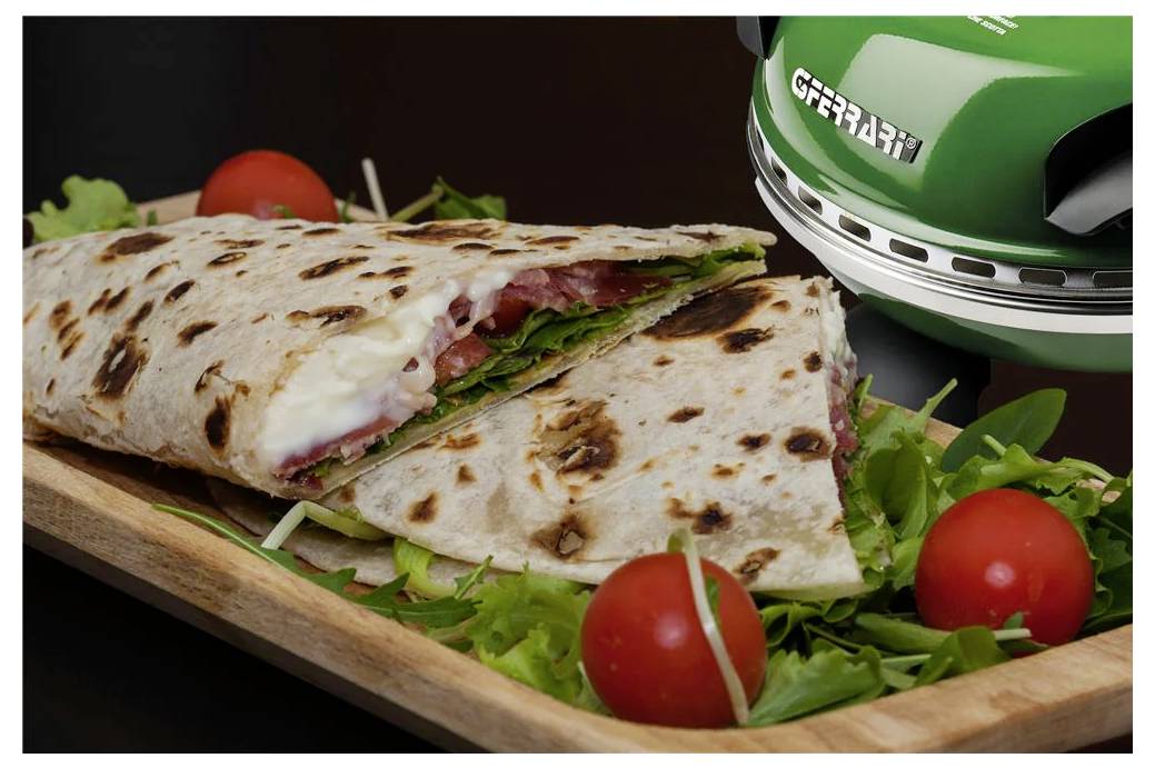 Grilovaný wrap sendvič nakrájený na polovinu na dřevěné desce, naplněný rozpuštěným sýrem a zeleninou, s rajčátky kolem zeleného panini lisu.