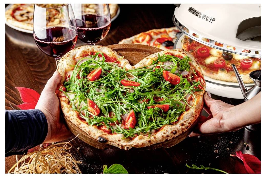 Dvě ruce podávají pizzu se zakysanou salátem a cherry rajčaty vedle sklenic červeného vína a stolní pece na pizzu.