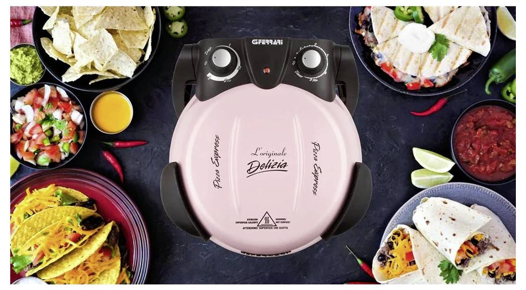Růžový elektrický pizzovací stroj Ferrari G3 umístěný uprostřed stolu, obklopený připravenými ingrediencemi a podávanými tacos a chipsy.