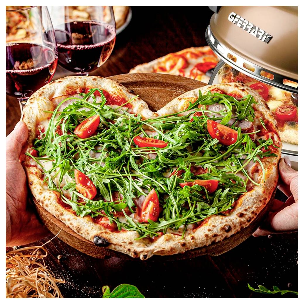 Pizza ve tvaru srdce s rukolou a cherry rajčaty, kterou někdo drží, v pozadí sklenice na víno a stolní gril.