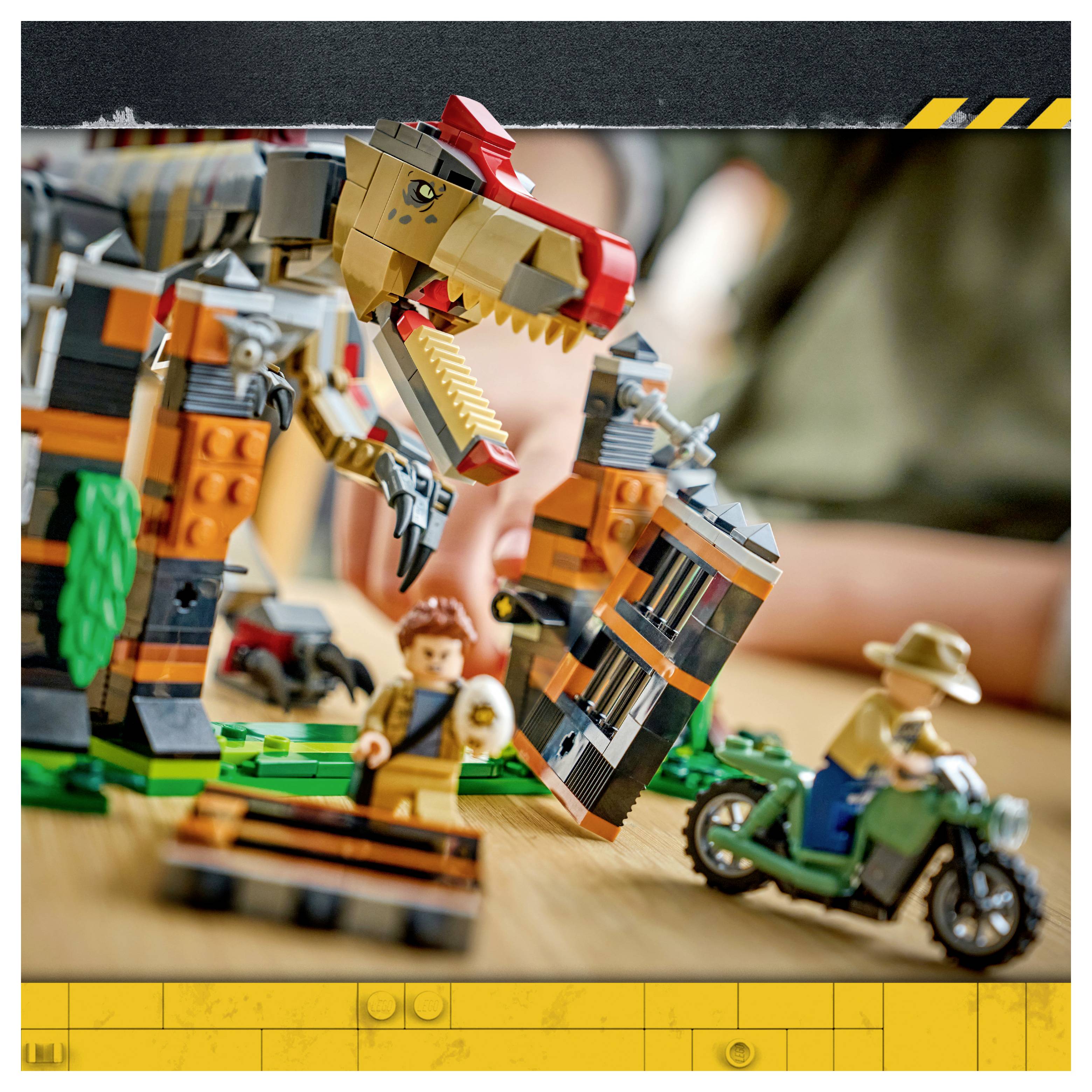 LEGO scéna s dinosaury se obřím T. rexem, minifigurkami a jezdcem na motorce na stolním setu.