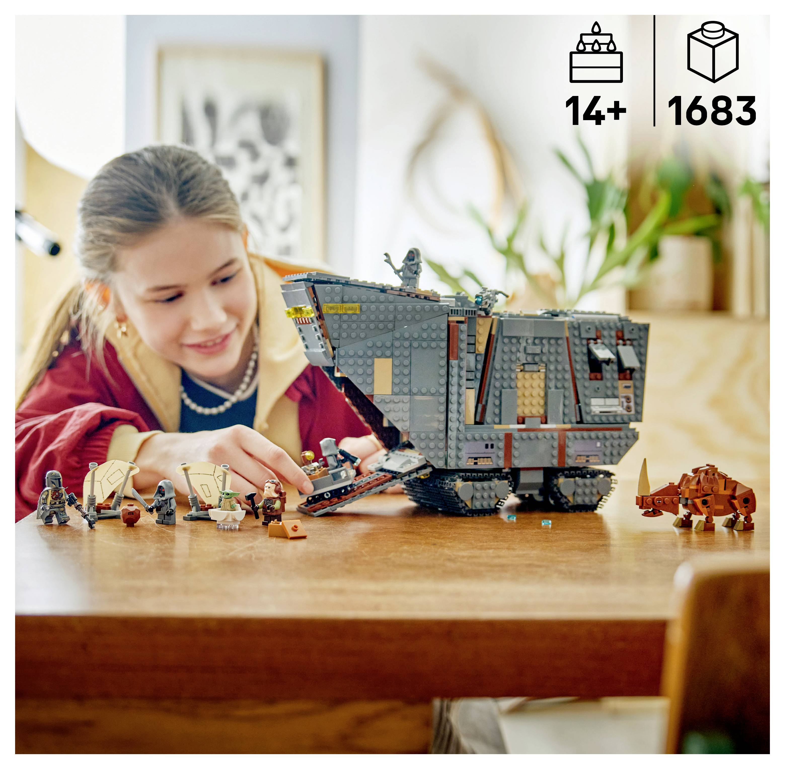 Dívka si hraje s velkým šedým modelem vesmírné lodi LEGO na stole, poblíž kterého je několik malých figurek a oranžové vozidlo.