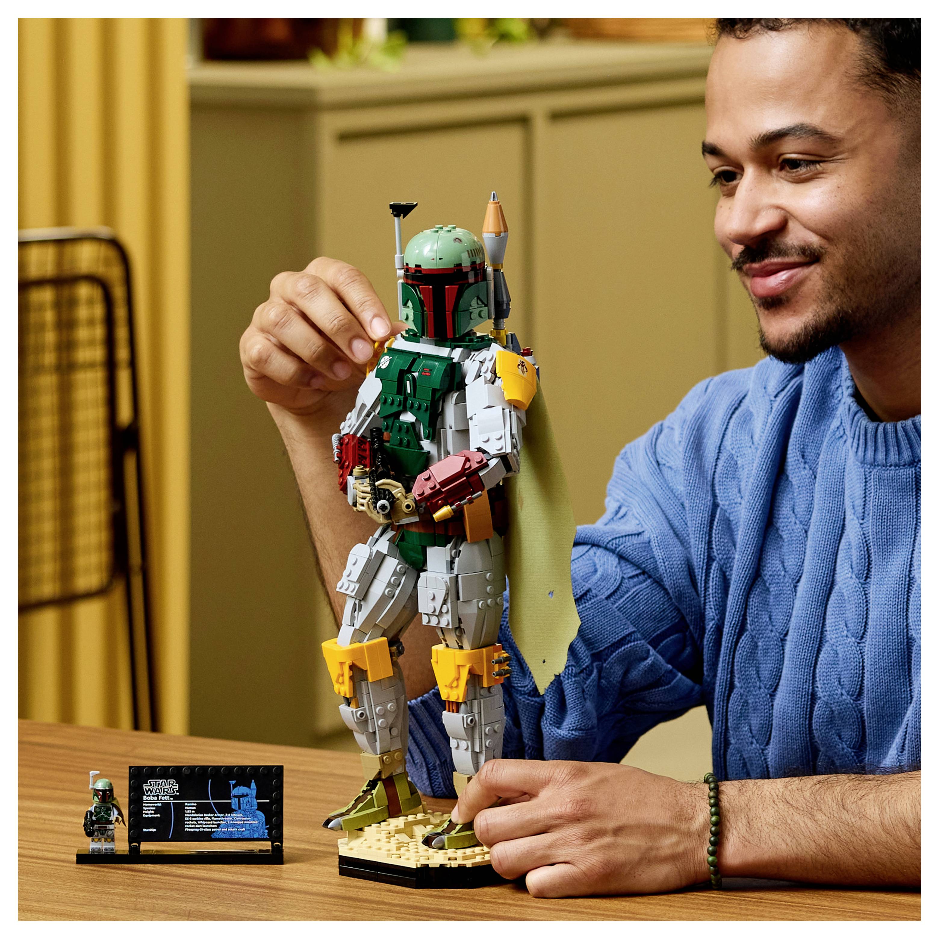 Muž v modré košili vystavuje LEGO figurku Boby Fetta na stole a drží ji vzpřímeně vedle její informační cedulky.