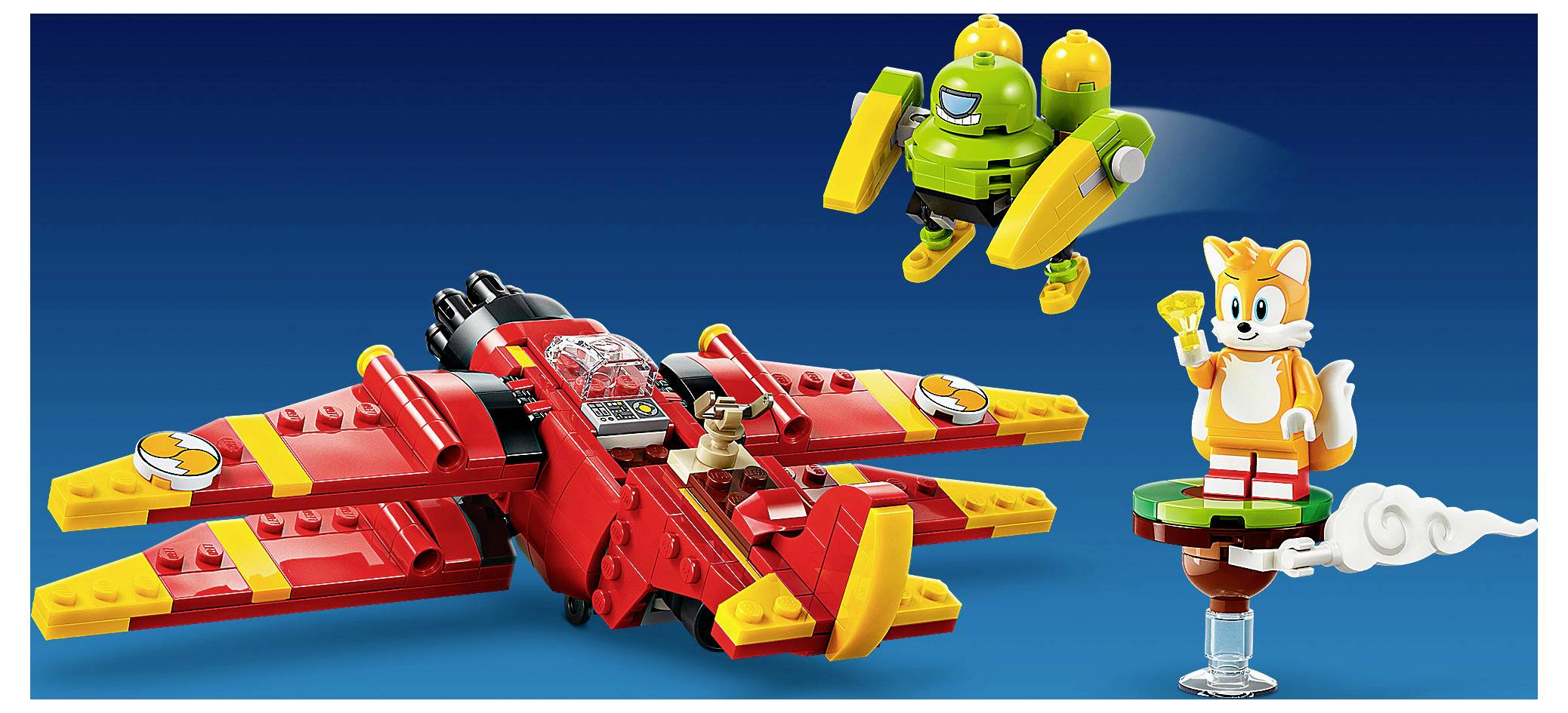 LEGO-stylový červený letoun s pilotem, zelený robot letící nad ním a Tails stojící na malé plošině, vše zobrazeno na modrém pozadí.