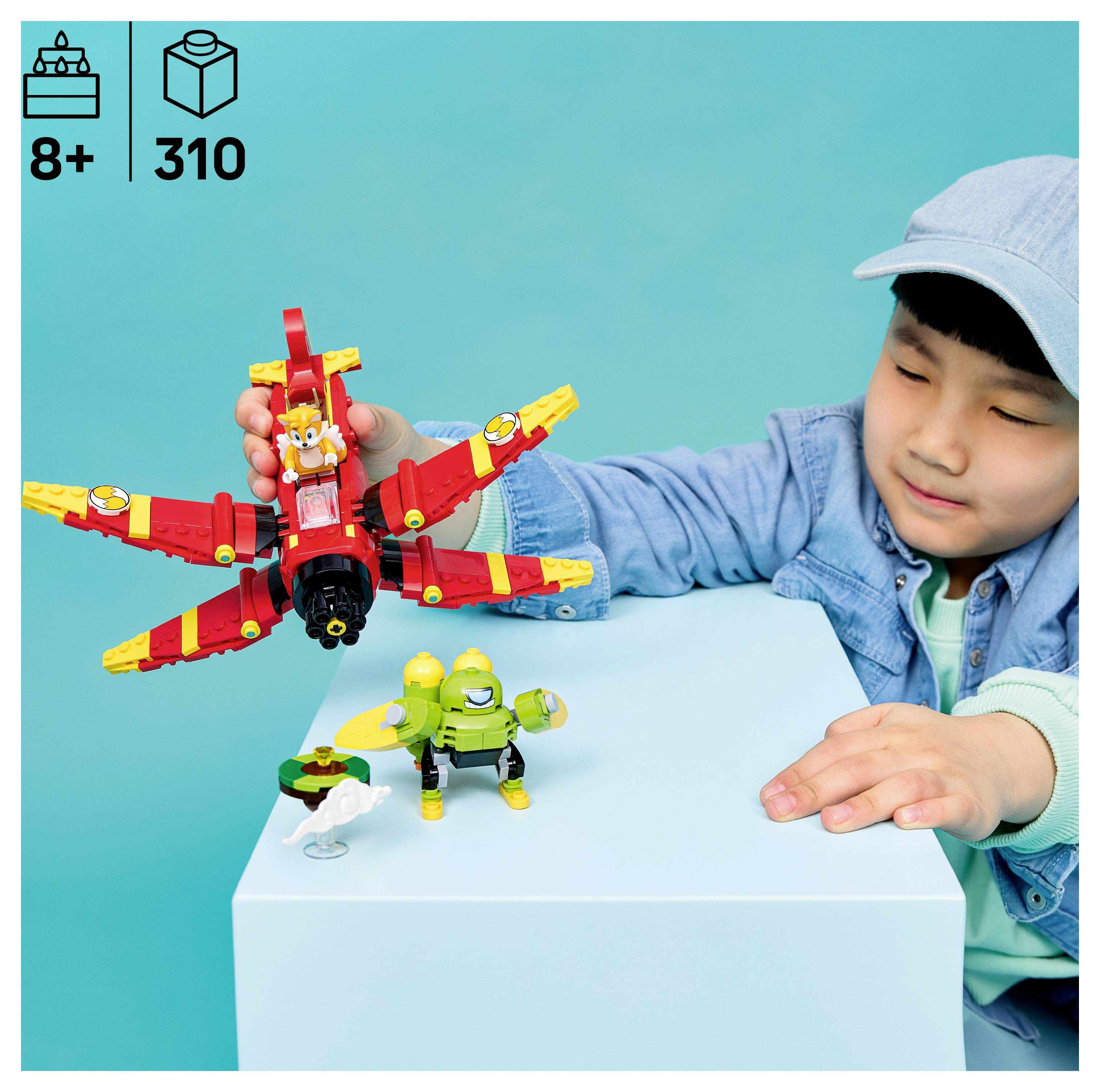 Mladý chlapec si hraje s červeno-žlutou LEGO létající bytostí a malými zelenými figurkami na bílém bloku, v rohu je vidět '8+' a '310'.