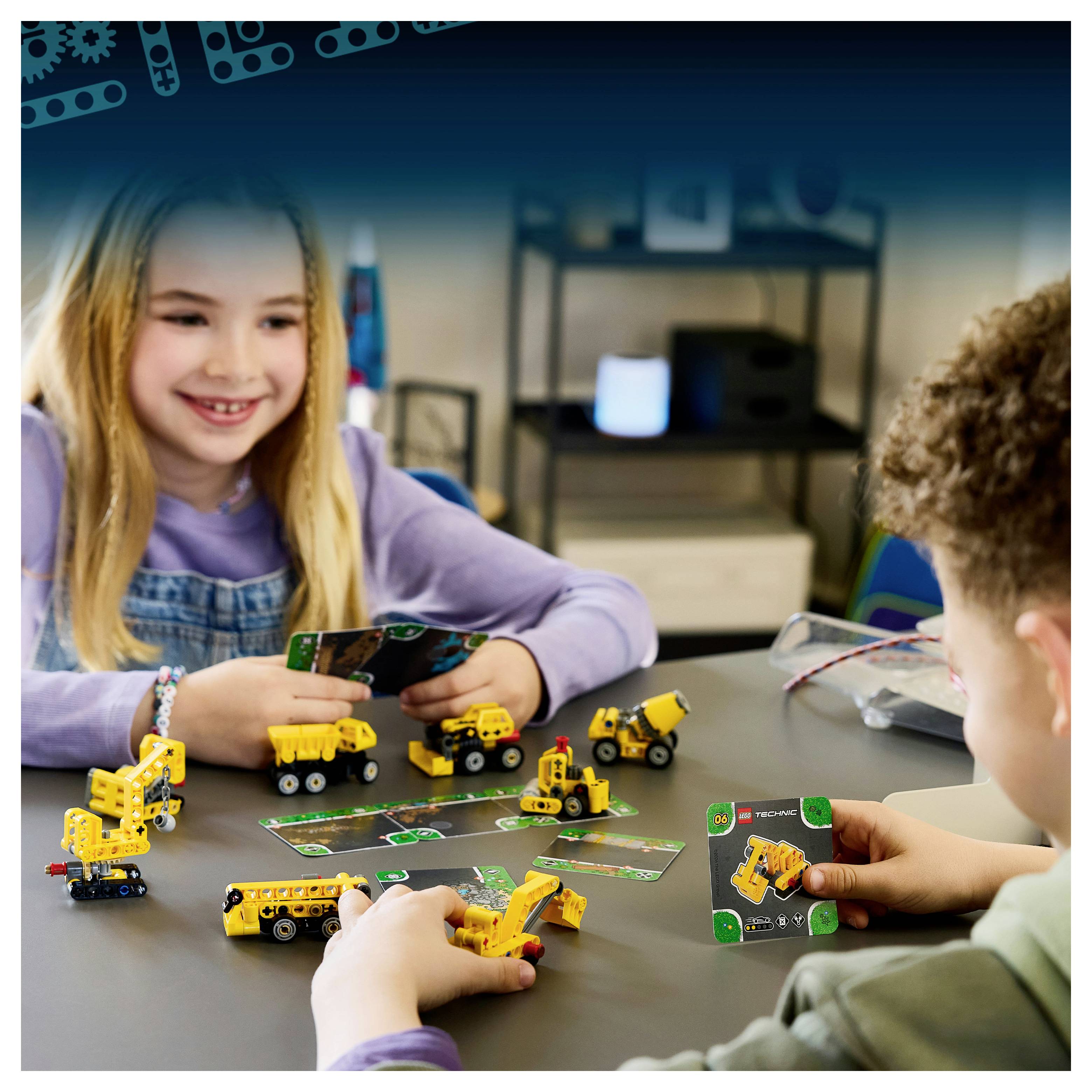 Dva děti si hrají s malými žlutými hračkami stavebních vozidel a hrními kartami u stolu ve třídě nebo herně.