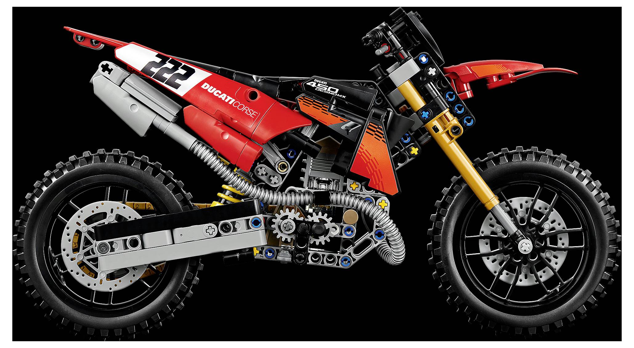 Boční pohled na červeném LEGO modelu motocyklu v stylu Ducati DesertX určeného pro off-road jízdu s viditelným motorem, odpružením a terénními pneumatikami na černém pozadí.