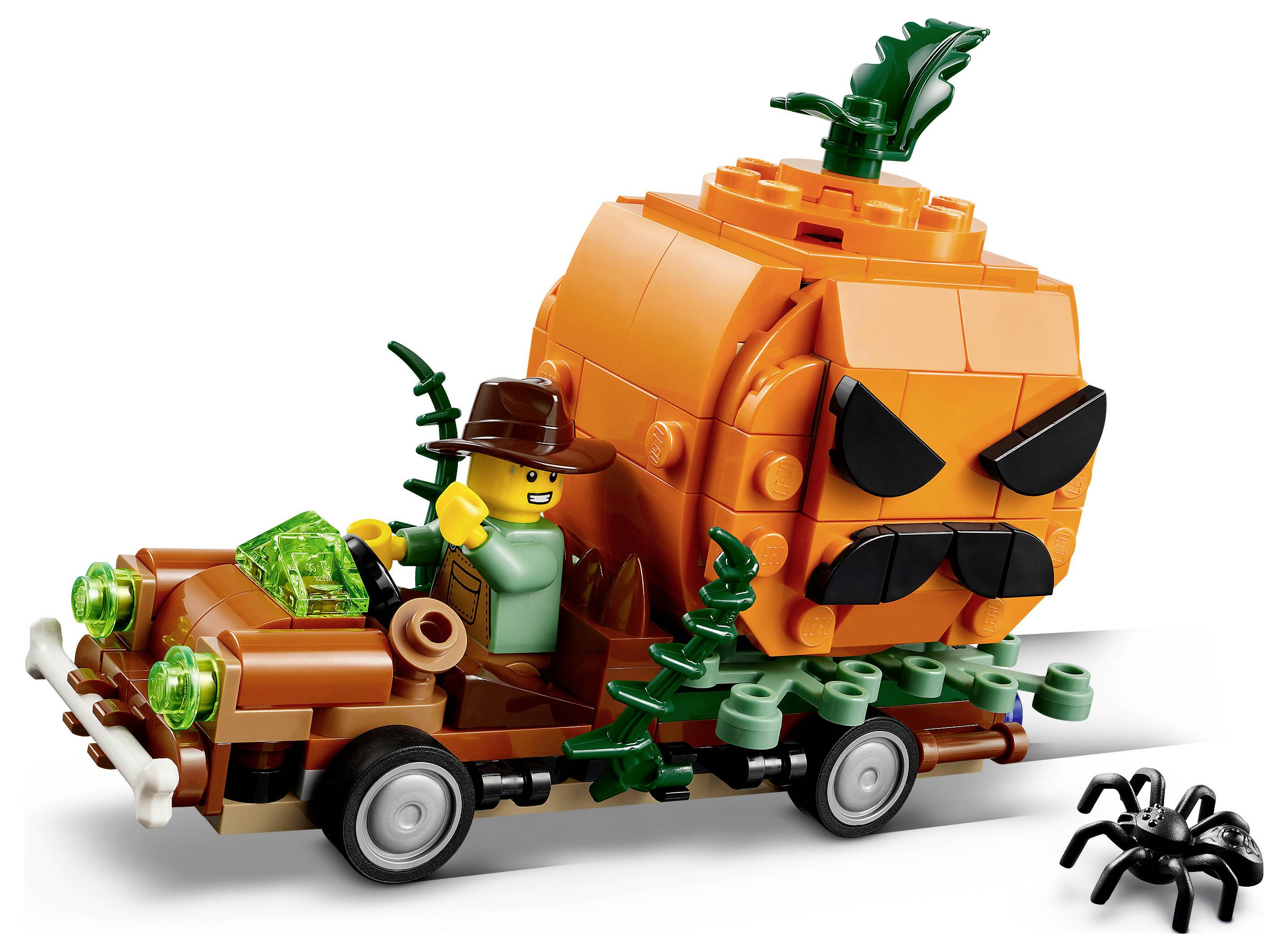 LEGO-style oranžový dýňový monster truck s řidičem v hnědém klobouku táhnoucí malý vozík se zelenými křišťálovými kousky; poblíž sedí černý pavouk.