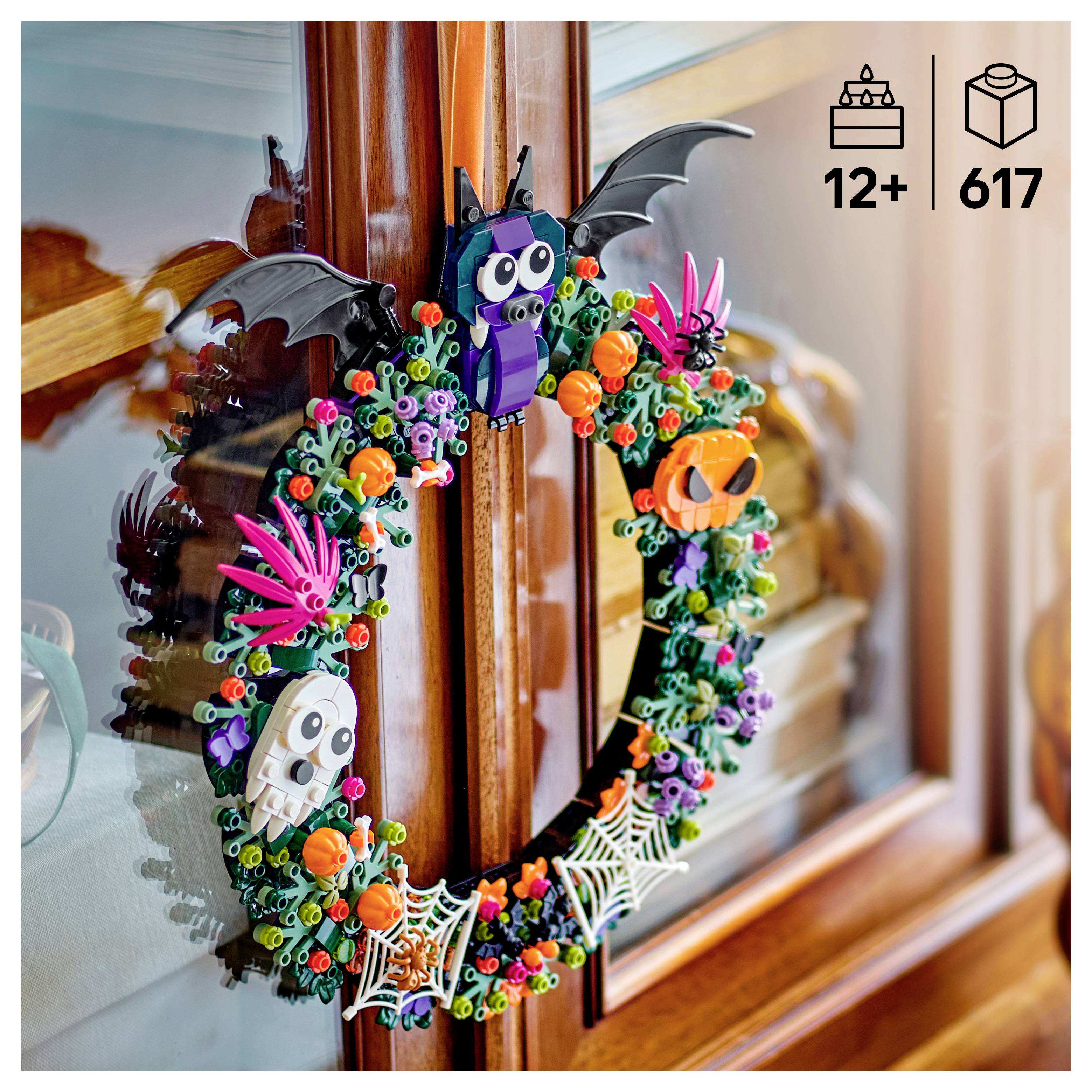 Barevný halloweenský věnec z LEGO s netopýry, lebkami, dýněmi, pavoučky a květinami visí na dřevěném okenním rámu.