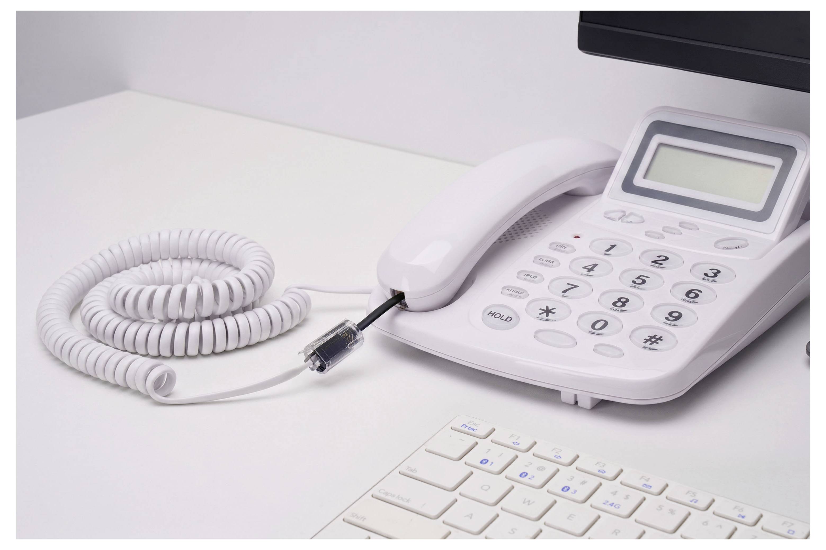 Bílý stolní kancelářský telefon s vinutým kabelem a sluchátkem spočívajícím na podstavci vedle klávesnice a monitoru.