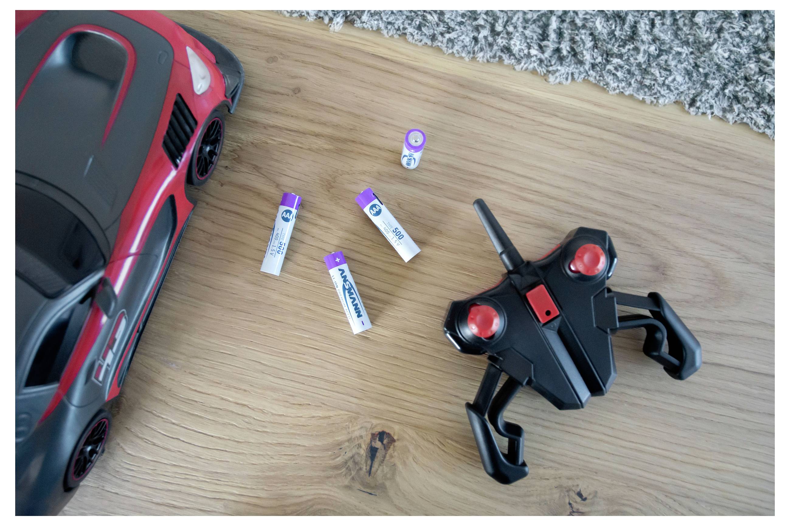 Červené hračkové auto a černý dálkový ovladač vedle několika baterií AAA na dřevěné podlaze
