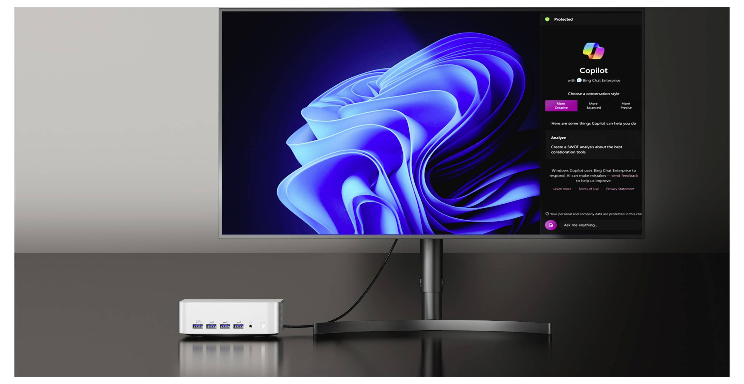 Stolní monitor zobrazuje rozhraní Windows 11 Copilot vedle modré tapety Windows, s malým dokovacím hubem na stole níže.