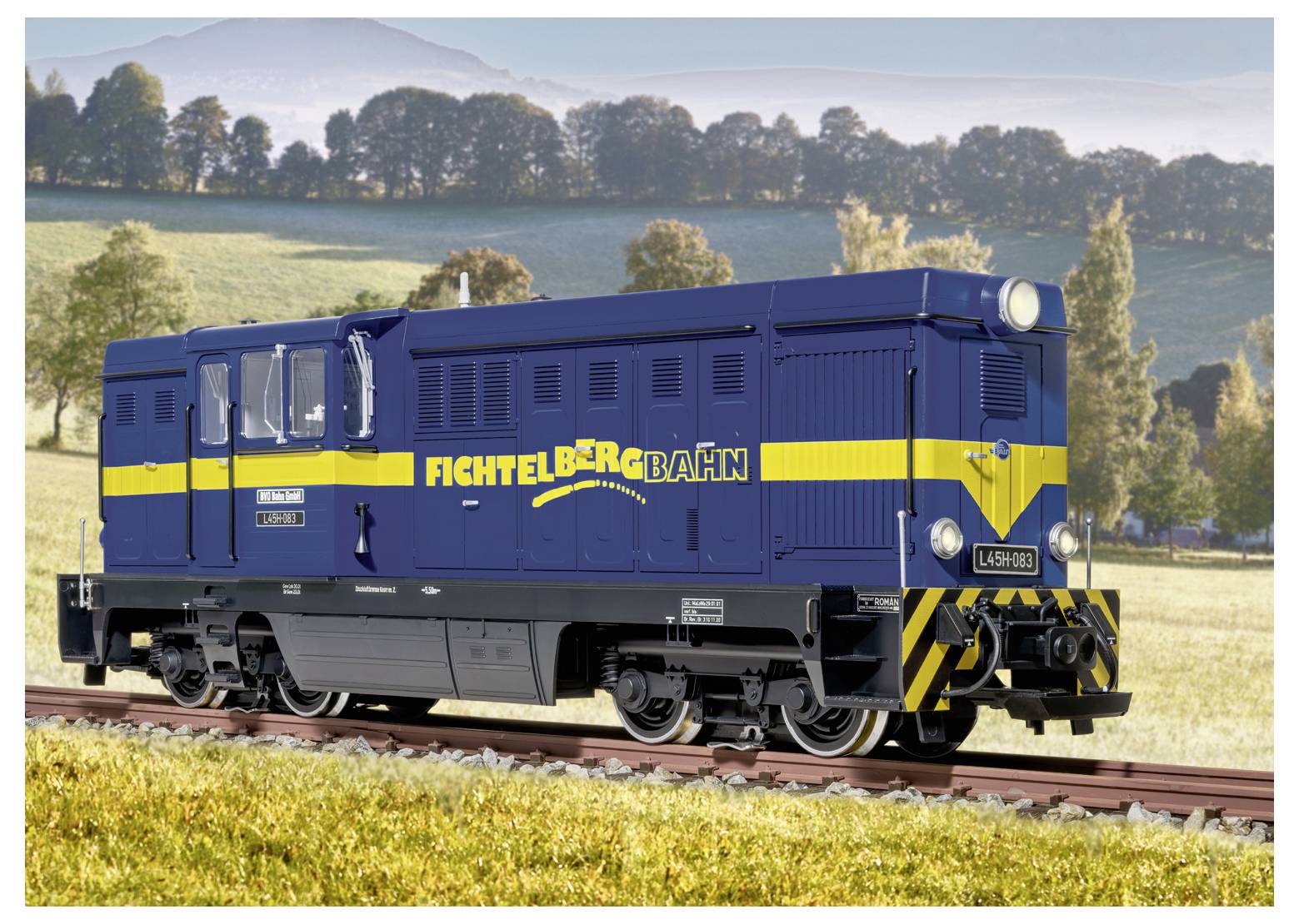 Modrá a žlutá dieselová lokomotiva Fichtelbergbahn na železničních kolejích v trávnaté venkovské krajině s kopci a stromy v pozadí.