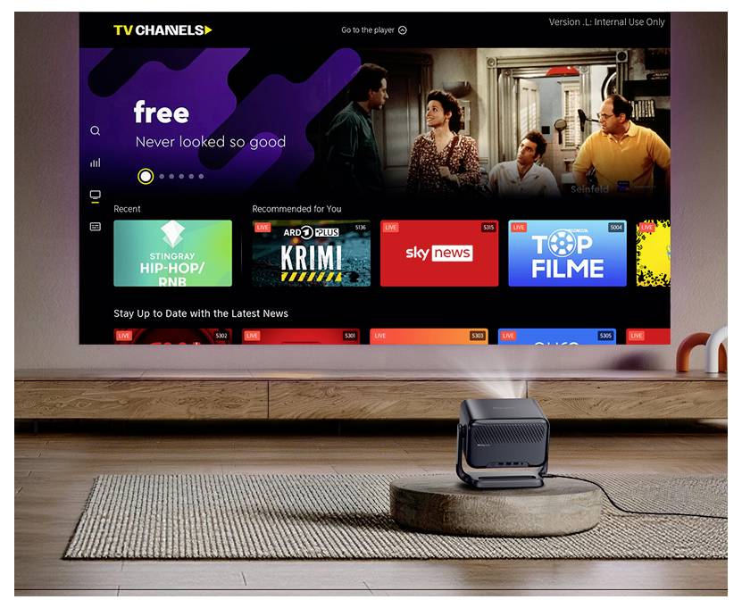 Domovská obrazovka Smart TV zobrazující bezplatné TV kanály a doporučené dlaždice obsahu, s bytovou scénou a zařízením podobným projektoru v popředí.