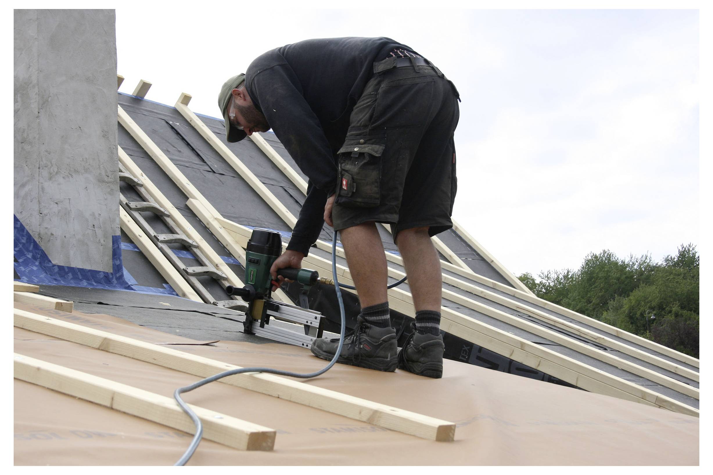 Pracovník používající pneumatickou hřebíkovnu k instalaci střešních desek na šikmé střeše během stavby.