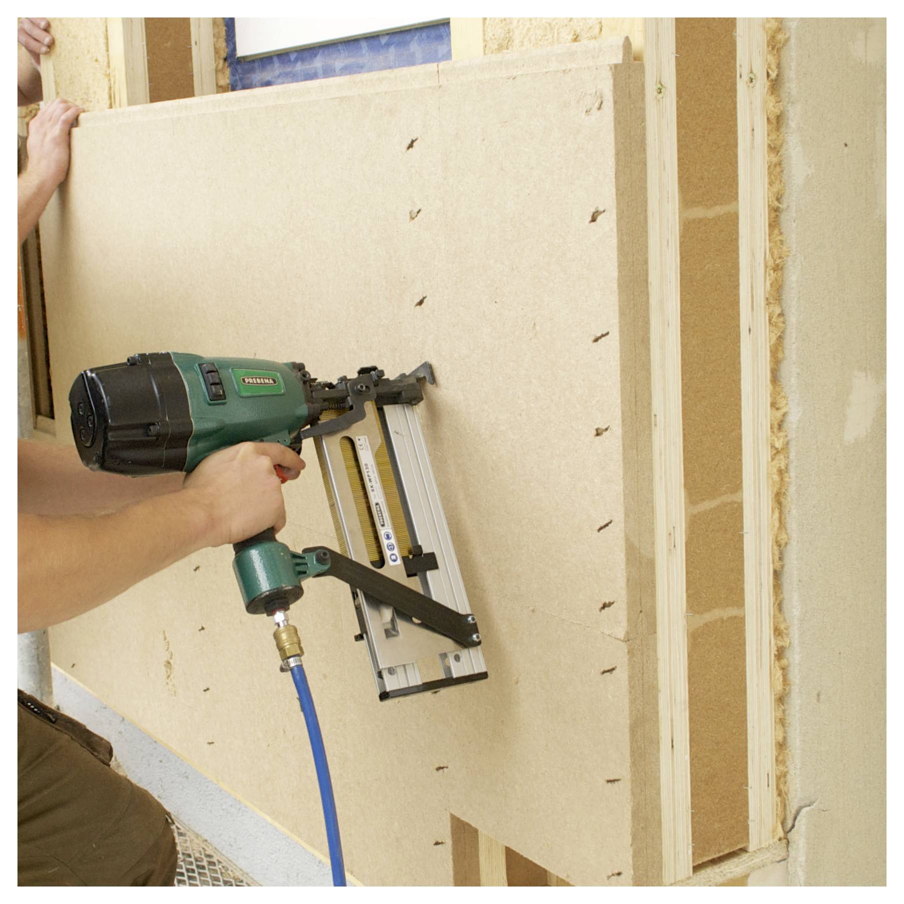 Pracovník upevňující velký béžový nástěnný panel pneumatickou hřebíkovnicí během stavby nebo renovace.