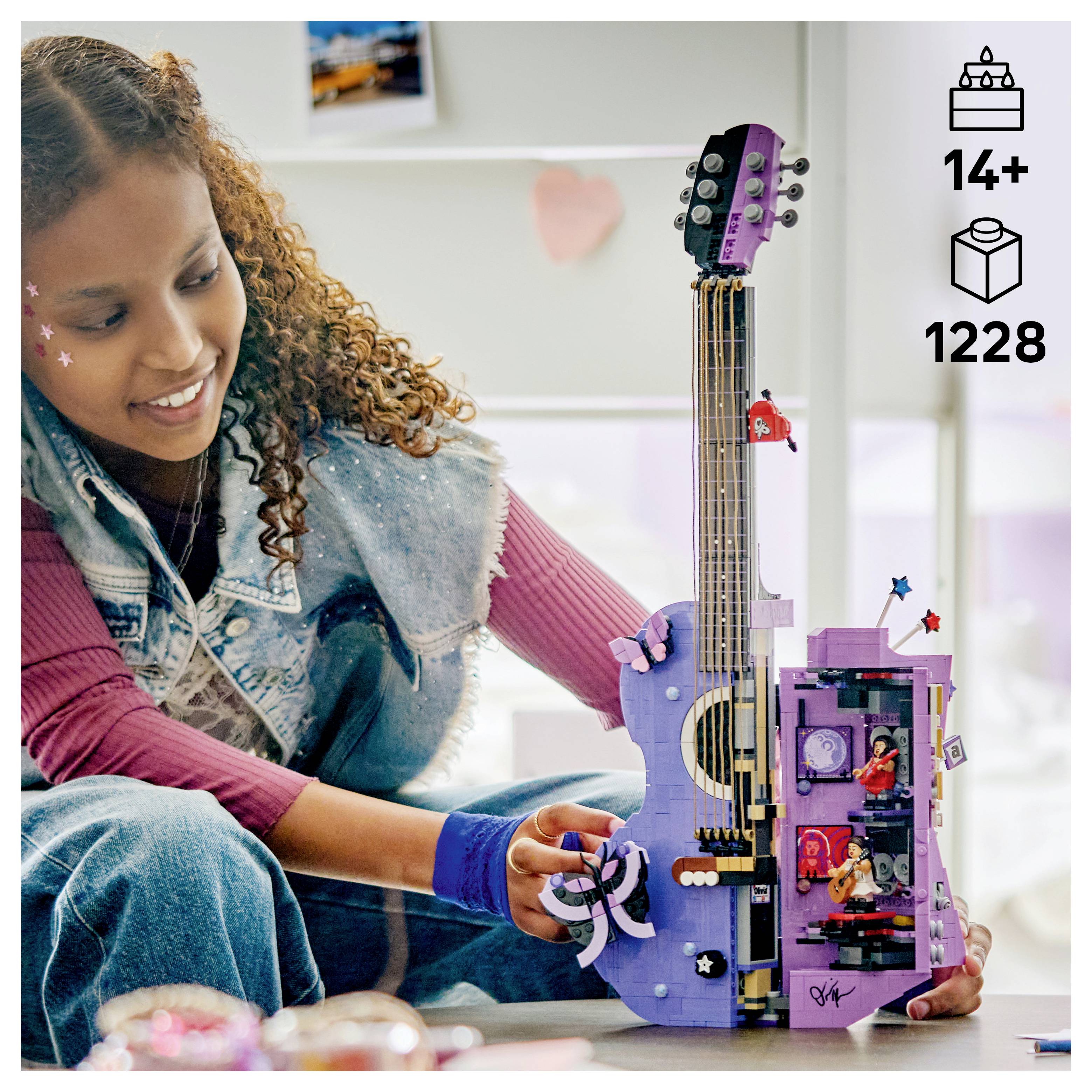 Dívka staví sadu fialové elektrické kytary LEGO, s ikonou věku 14+ a 1228 kusů zobrazenými na krabici.