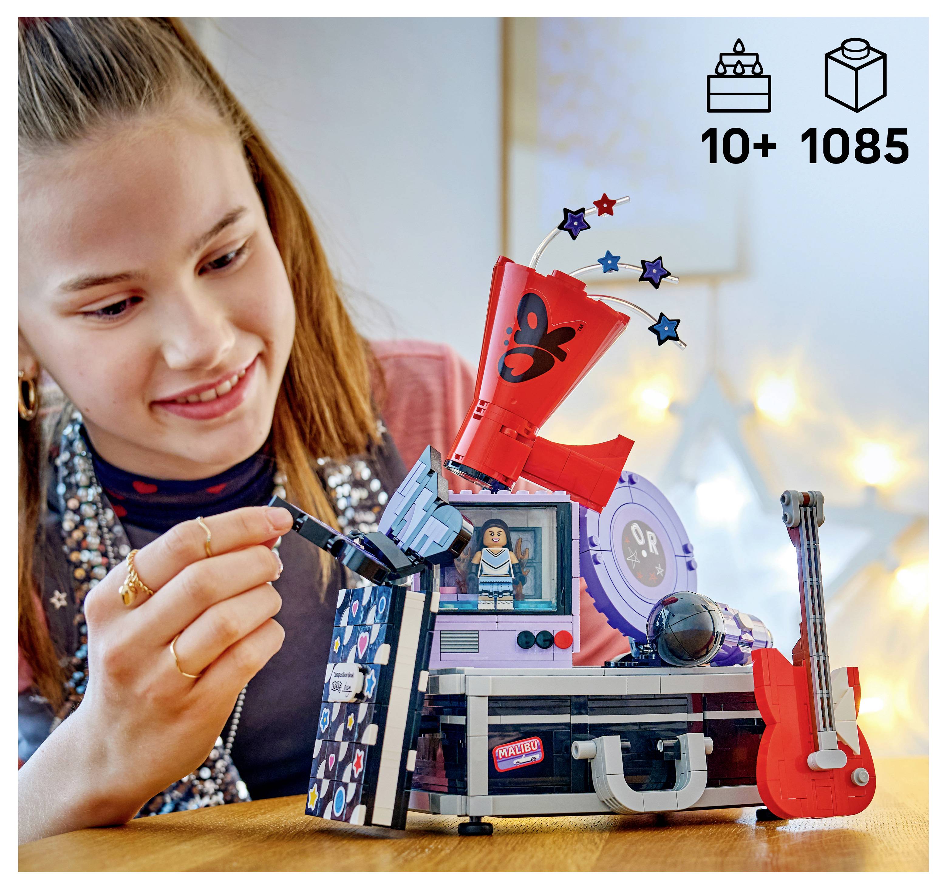 Dívka si skládá LEGO sadu s hudební tématikou s červenou kytarou, scénickými světly a minifigurkou; ikony ukazují 10+ let a 1085 součástek.