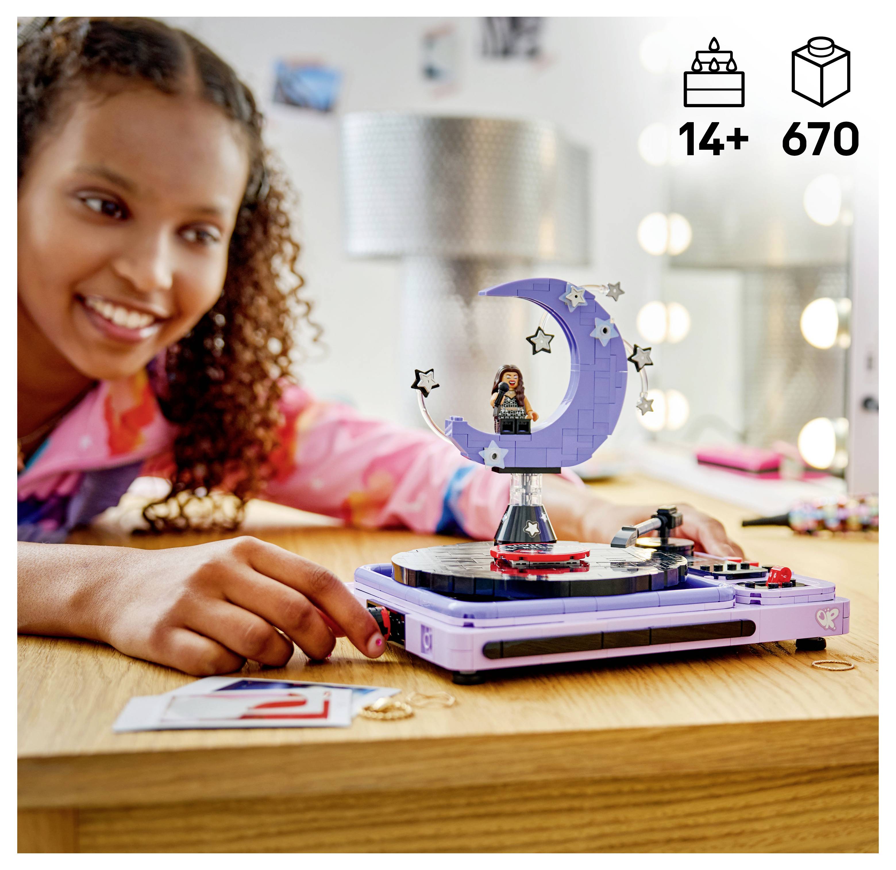 Usmívající se dívka vytváří displej měsíce ve stylu LEGO na otočném stole, s ikonami „14+" a „670" dílků v rohu.