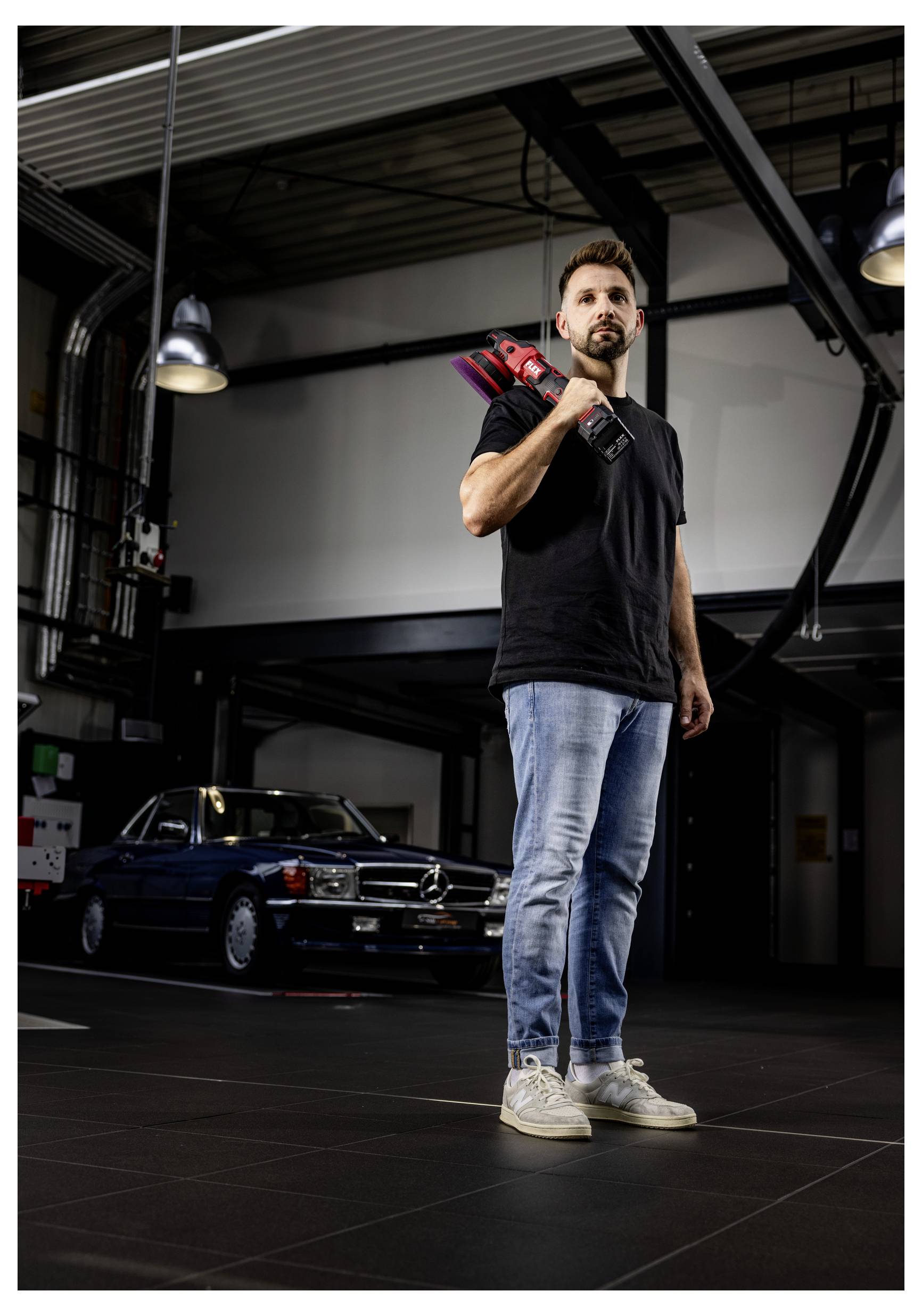 Muž drží v garáži červený elektrický nástroj přes rameno, zatímco stojí před klasickým černým automobilem.