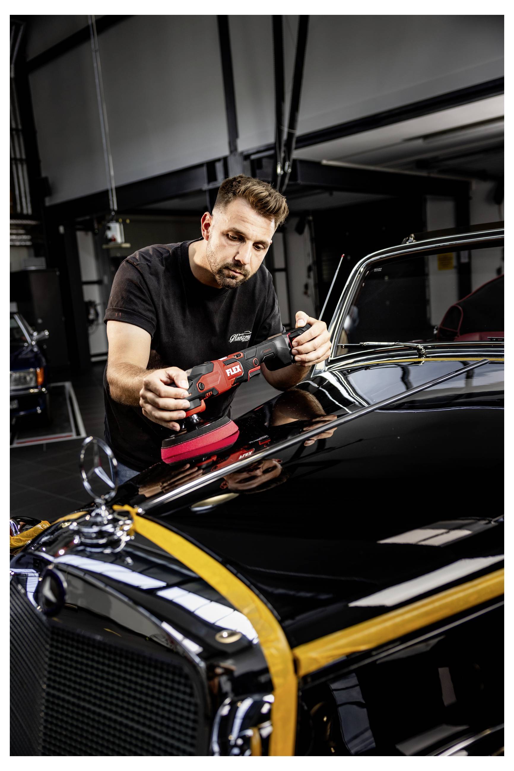 Muž leštící černý klasický automobil ruční leštičkou v garáži, zaměřený na kapotu a chromované obložení.